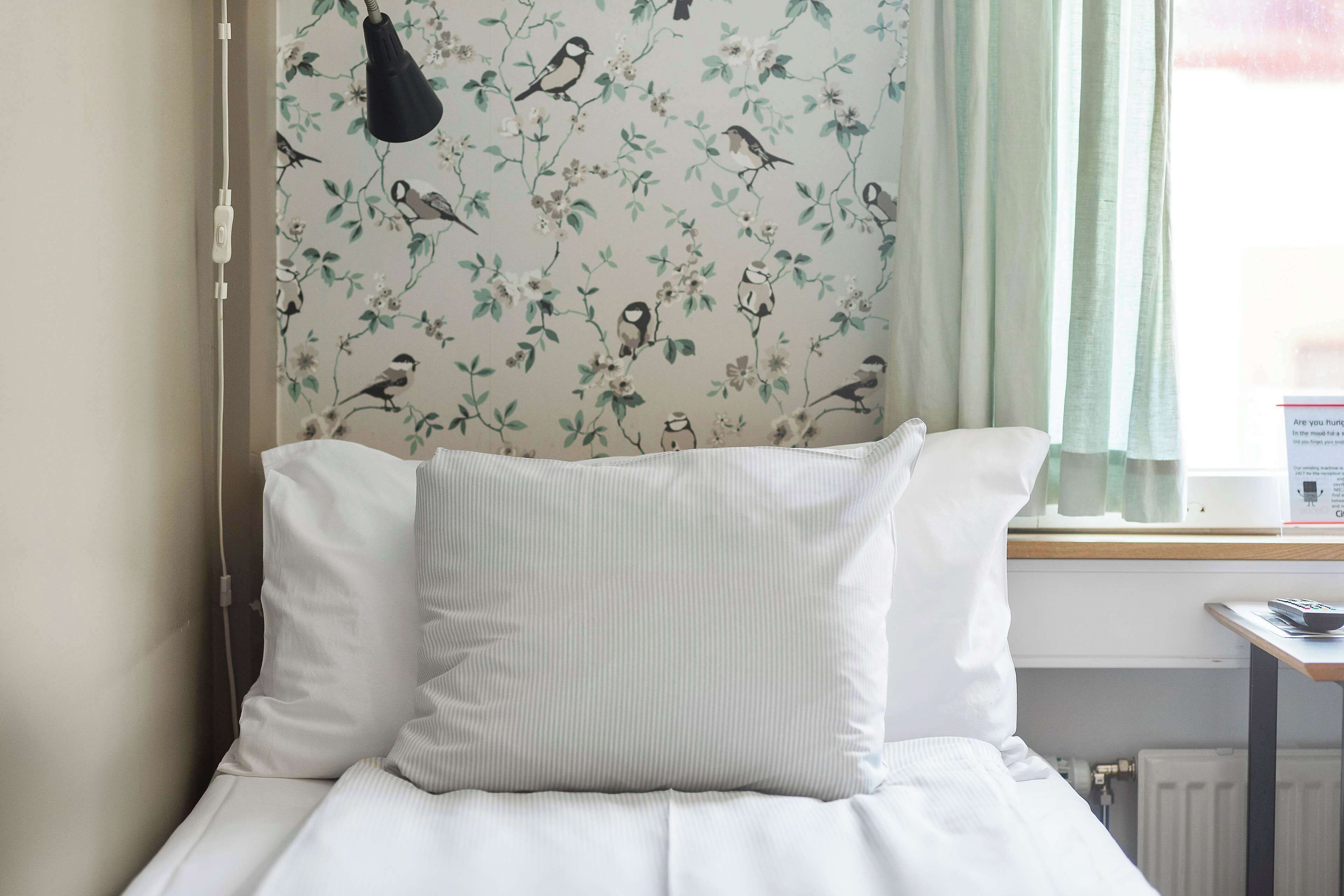 Una cama con ropa blanca y una almohada, una ventana con vista a pájaros en la pared, y una lámpara en la mesita de noche.
