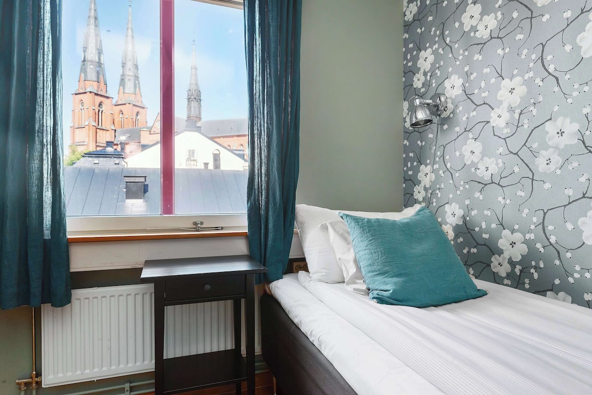 A bedroom with a bed, a window with curtains, a radiator, and a wall with a floral pattern.
