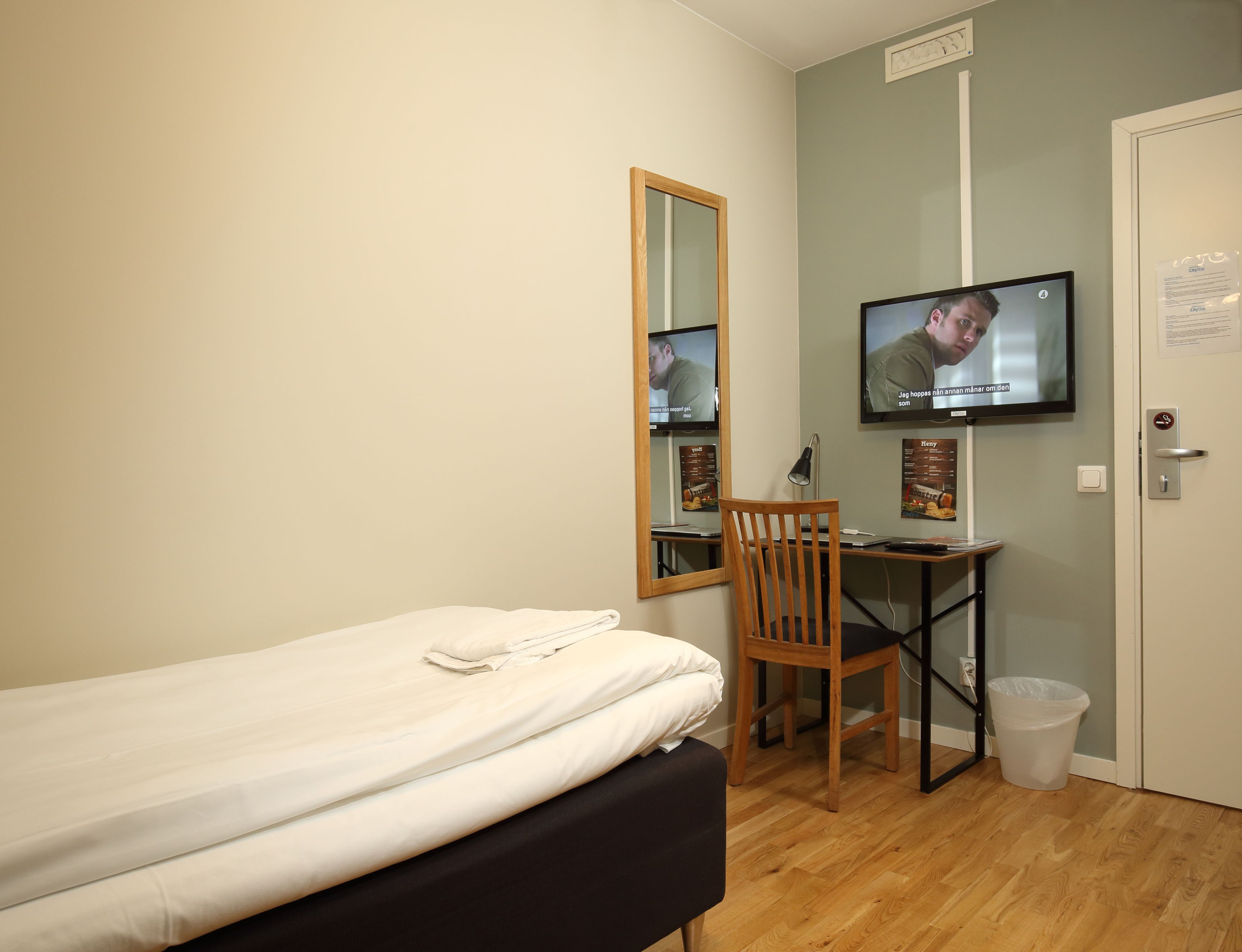A small room with a bed, a desk, a chair, a TV, and a trash bin.