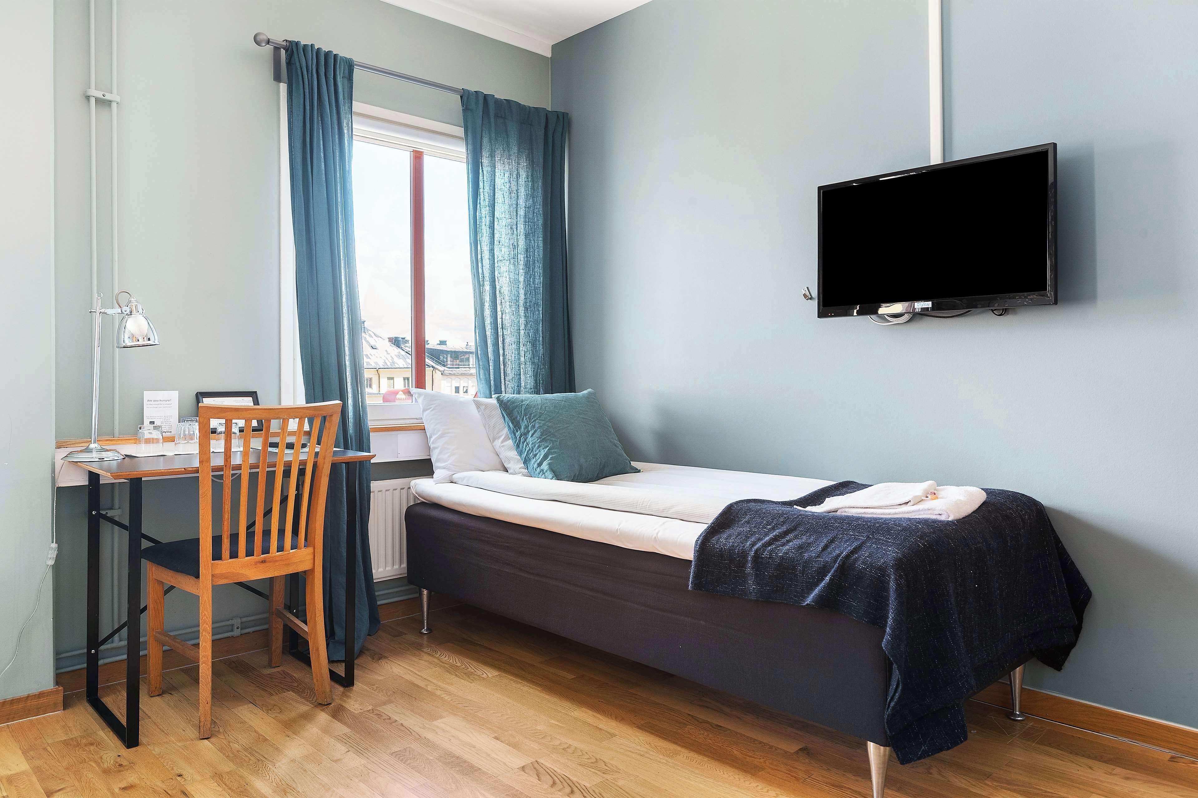 Une petite pièce comprenant un lit, un bureau, une chaise, un téléviseur et une fenêtre avec des rideaux.