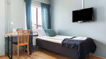 Une petite pièce comprenant un lit, un bureau, une chaise, un téléviseur et une fenêtre avec des rideaux.