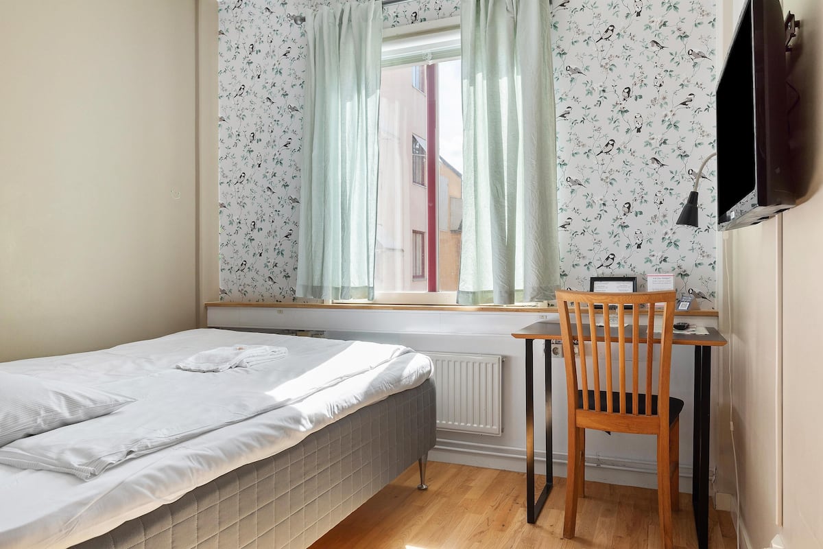 A bedroom with a single bed, a window with curtains, a desk with a chair, and a radiator.