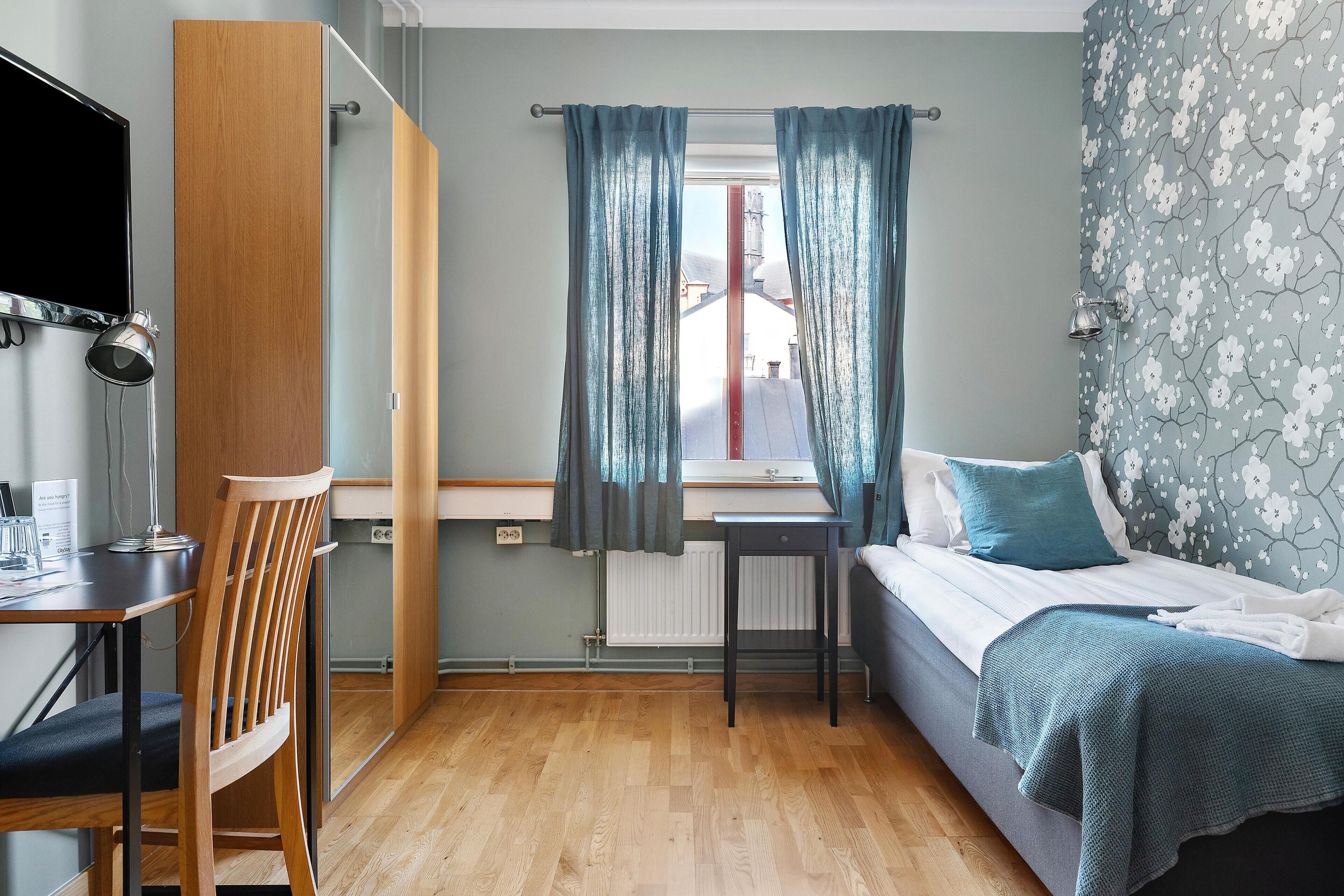A bedroom with a bed, a desk, a chair, and a window with curtains.