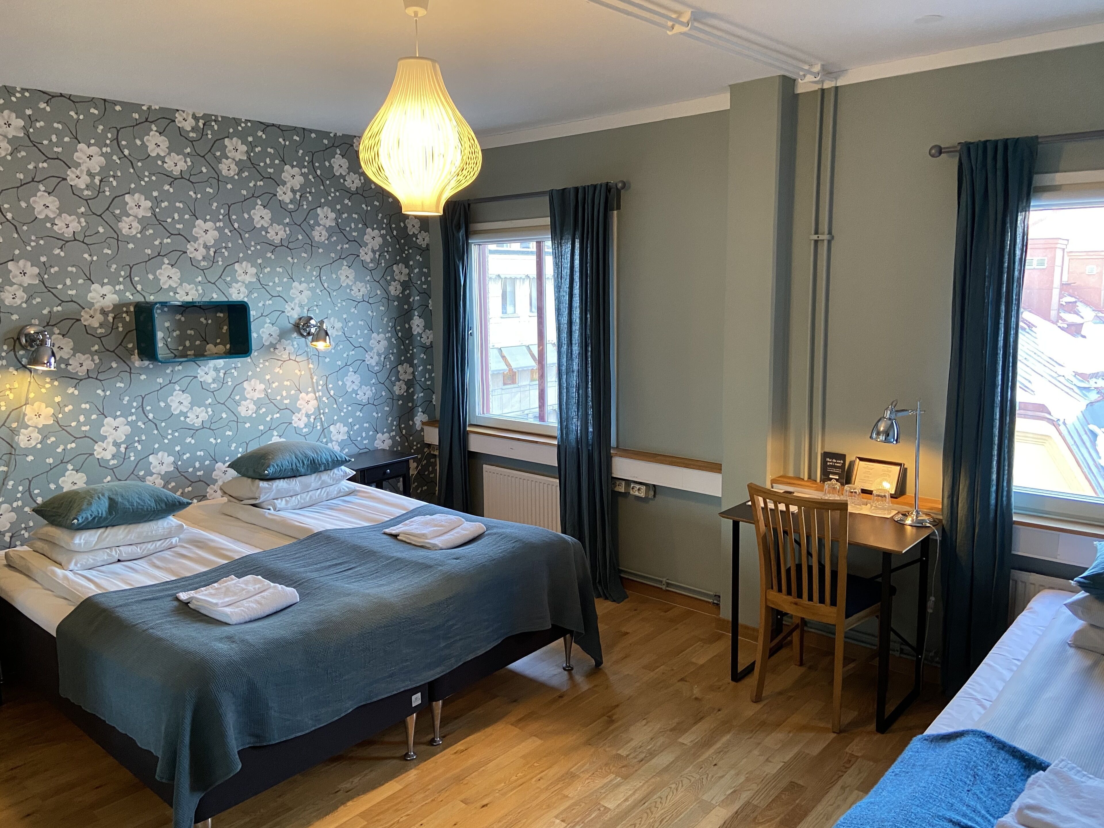 Une chambre d’hôtel avec un lit double, un bureau, une chaise, une fenêtre avec des rideaux, une lampe fixée au mur et un panneau décoratif mural.