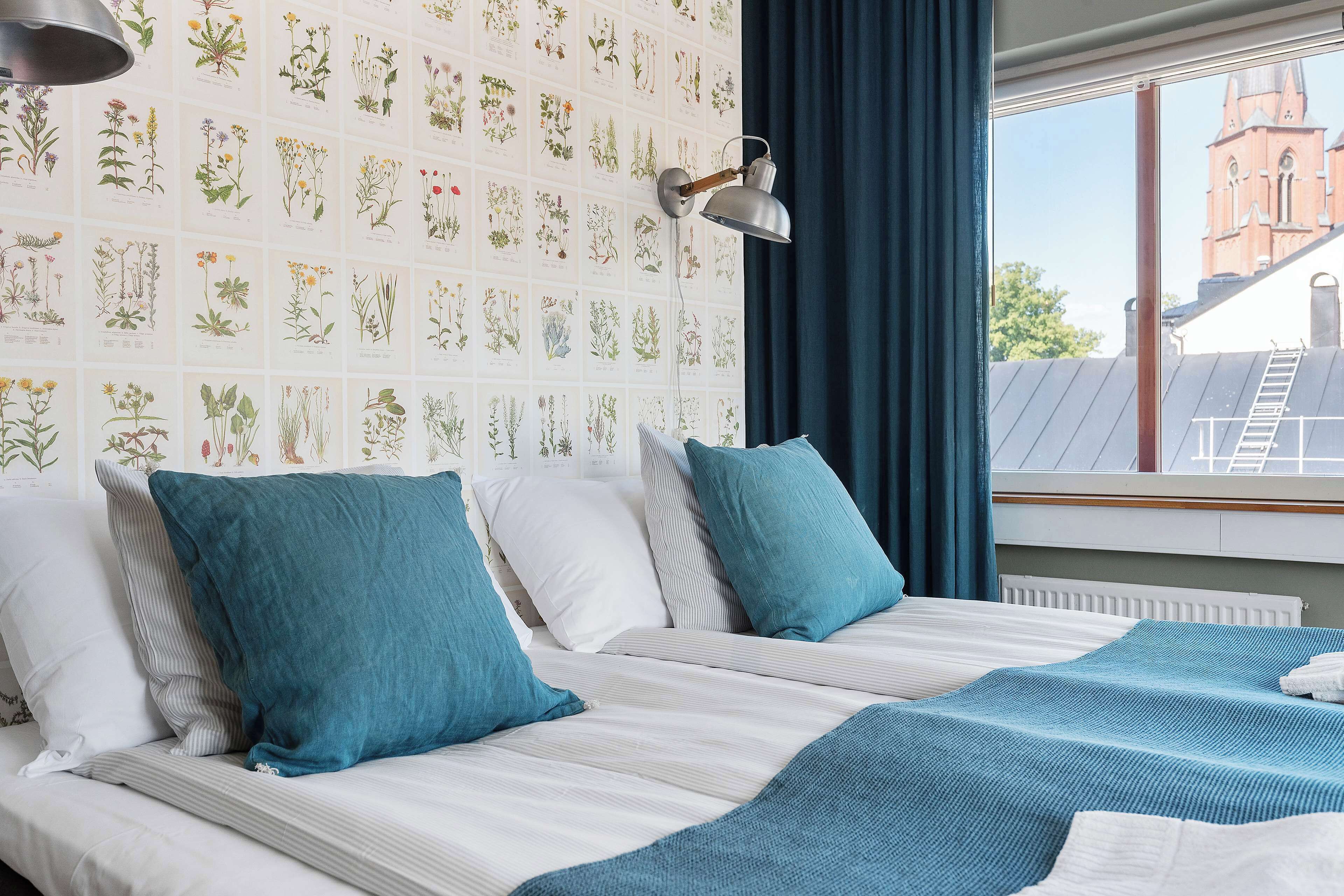 A hotel room with a large bed, two pillows, a blue blanket, a wall with botanical illustrations, a window with a view of a building, and a lamp.