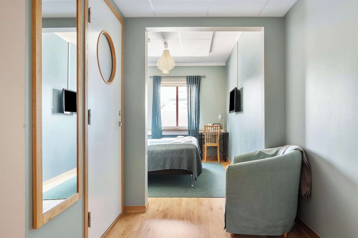 A narrow hallway leading to a bedroom with a bed, a chair, a desk, and a television.