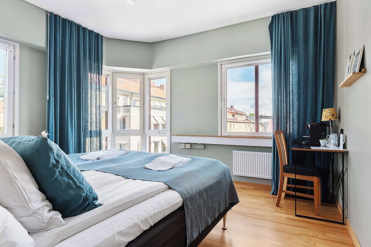 A bedroom with a bed, a desk, a chair, and a view of buildings through the window.