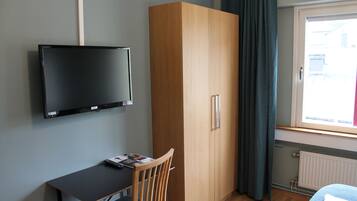 A room with a bed, a desk with a chair, a wardrobe, a television mounted on the wall, and a window with curtains.
