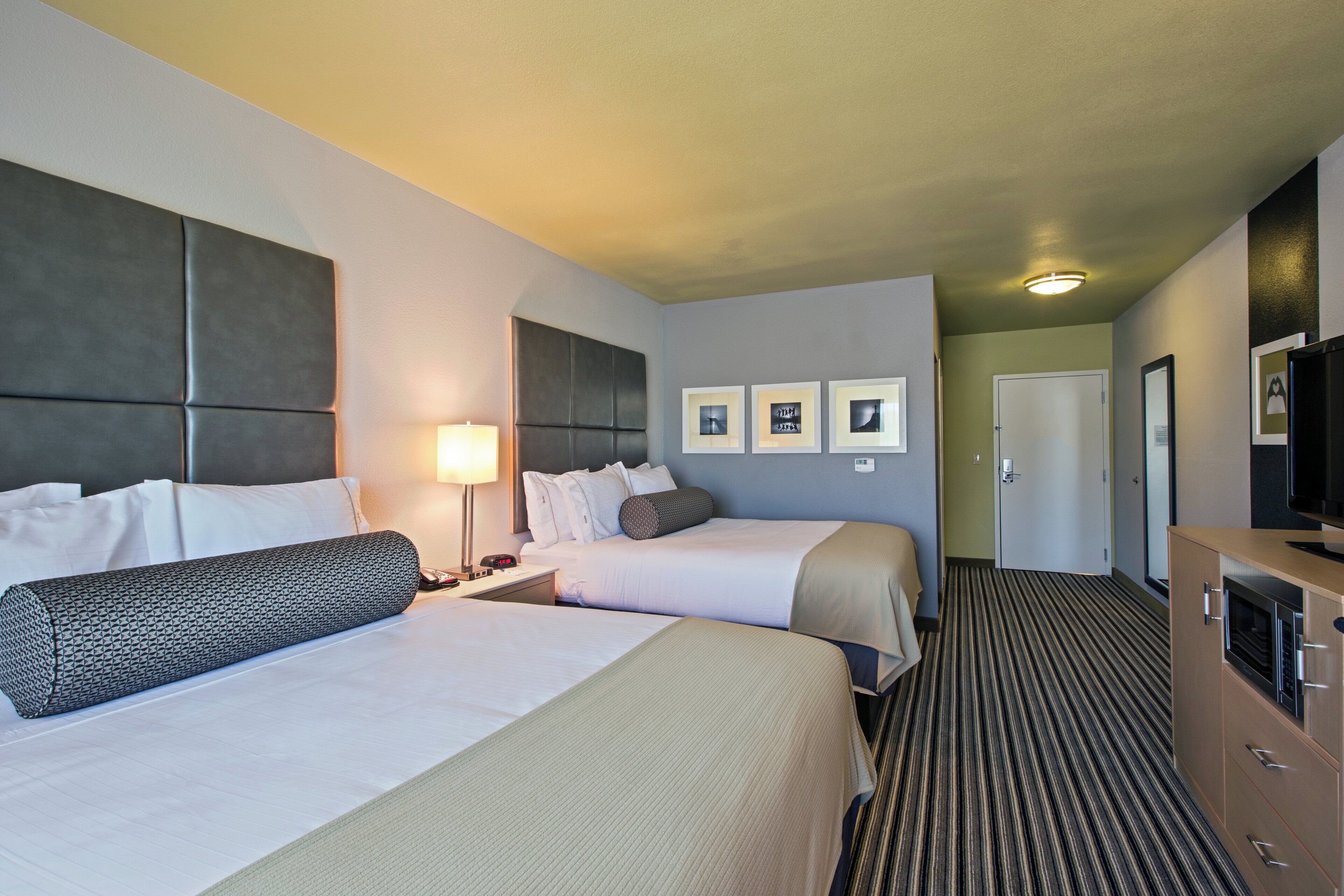 A hotel room with two beds, a bedside table with a lamp, a television, and a door leading to another room.