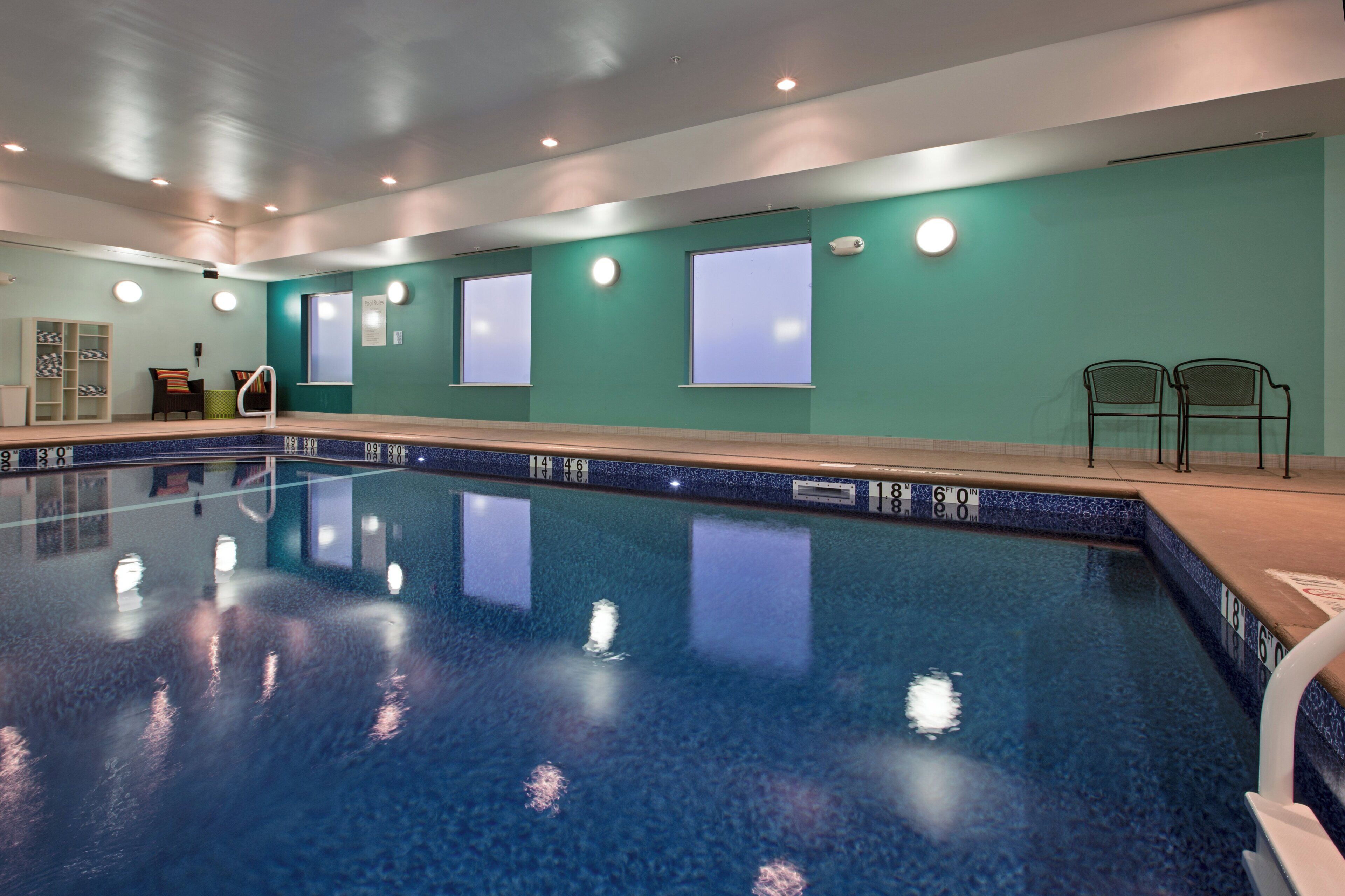 Una piscina con agua cristalina, rodeada por una solana empavedada y un muro verde con ventanas.