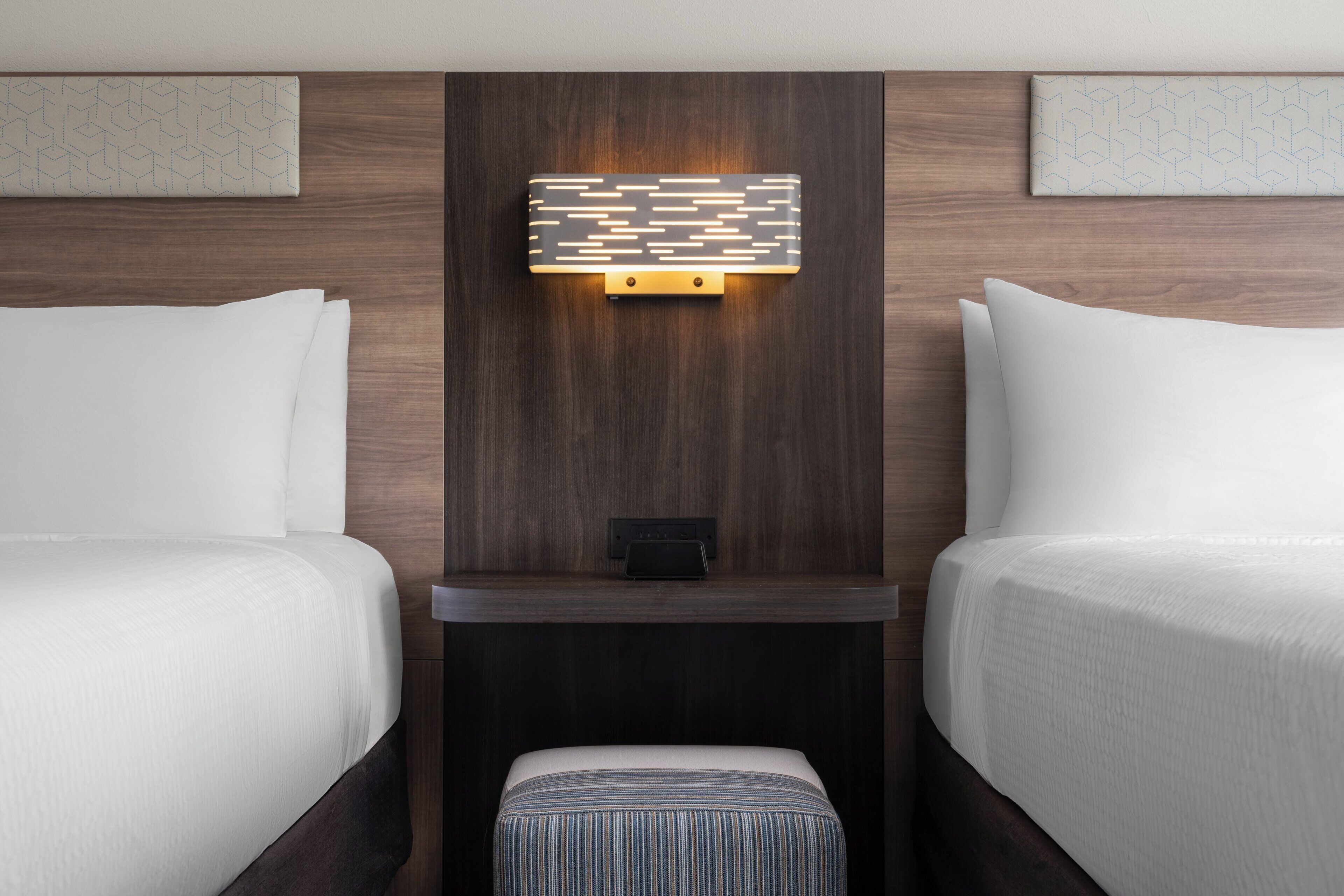 A hotel room with two beds, a nightstand, a wall-mounted light fixture, and a small table.