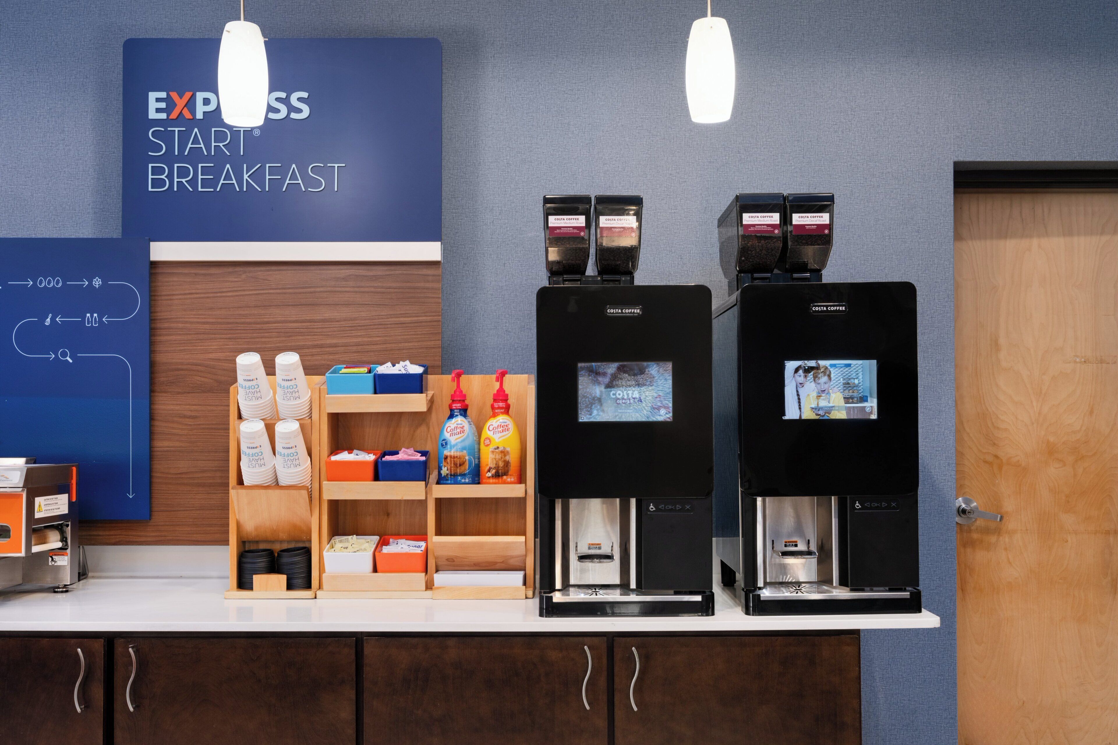 Um espaço para café com duas máquinas de café, uma placa com a inscrição 'EXPRESS START BREAKFAST' e uma seleção de cápsulas e garrafas de café.