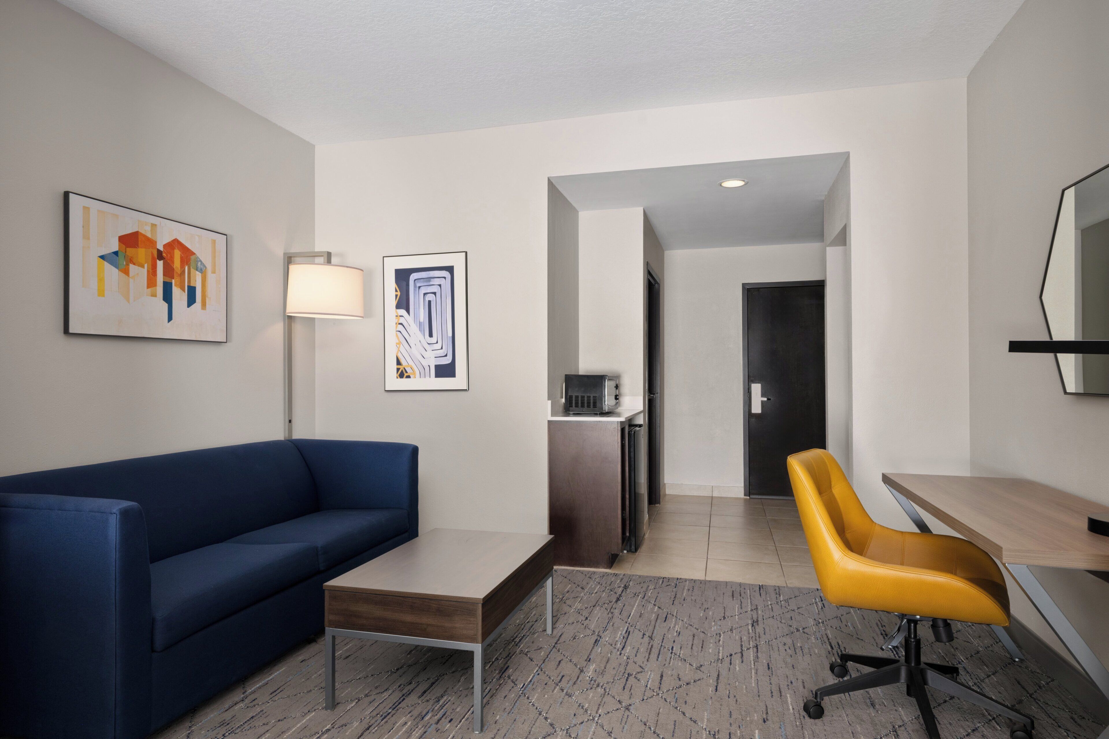 A modern hotel room with a blue sofa, a wooden desk, a yellow office chair, a standing lamp, and artwork on the walls.