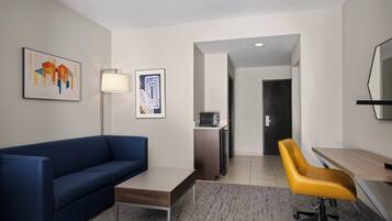 A modern hotel room with a blue sofa, a wooden desk, a yellow office chair, a standing lamp, and artwork on the walls.