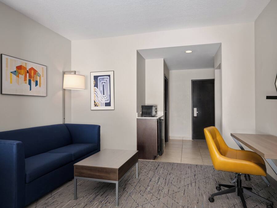 A modern hotel room with a blue sofa, a wooden desk, a yellow office chair, a standing lamp, and artwork on the walls.