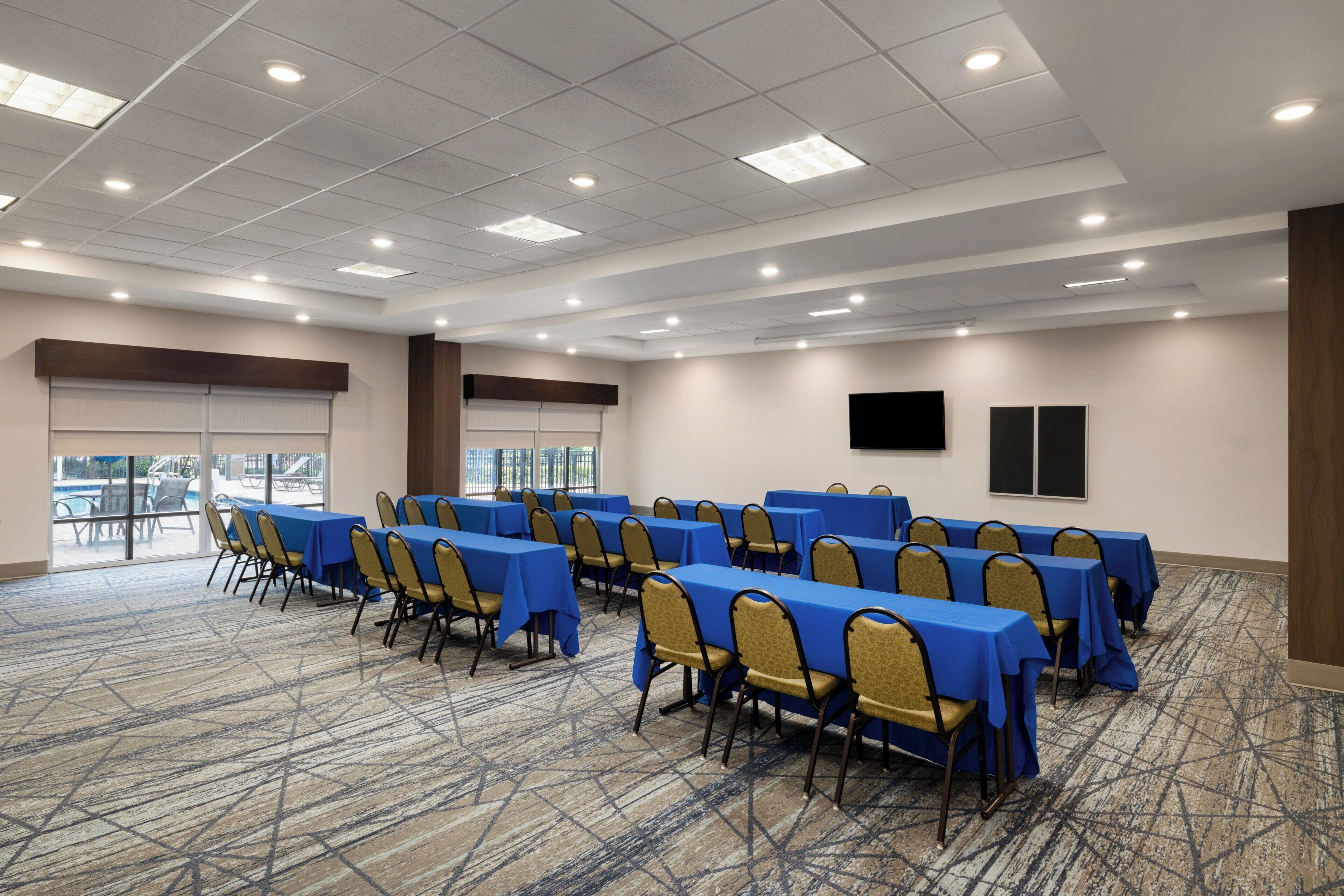 Une salle de conférence équipée de tables et de chaises, de grandes fenêtres et d’un sol recouvert de moquette.