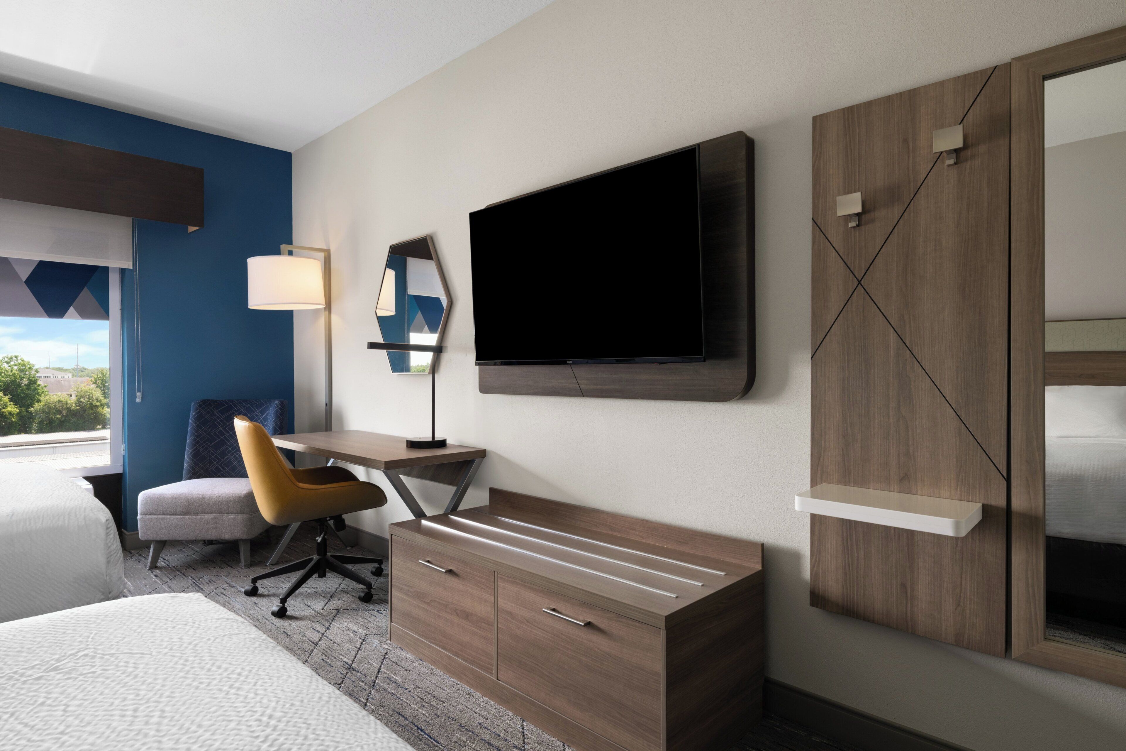A modern hotel room with a bed, a desk with a chair, a TV mounted on the wall, and a wardrobe.