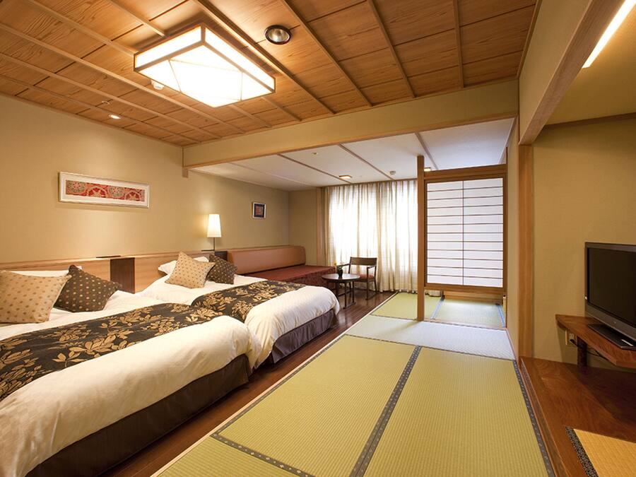 A hotel room with two beds, a television, tatami mats, a window with curtains, and a wooden ceiling.