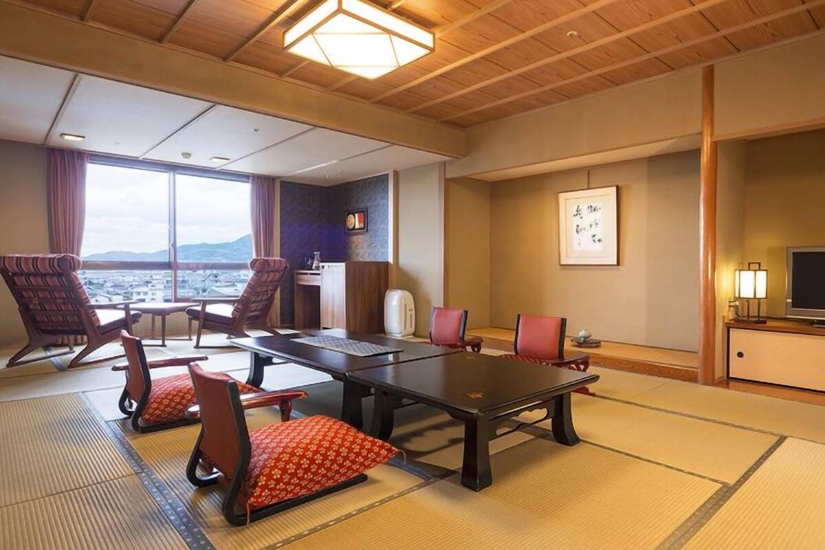 Une chambre d’hôtel avec des tatamis, une grande fenêtre donnant sur un beau paysage, une table à manger avec des chaises, un téléviseur et un meuble.
