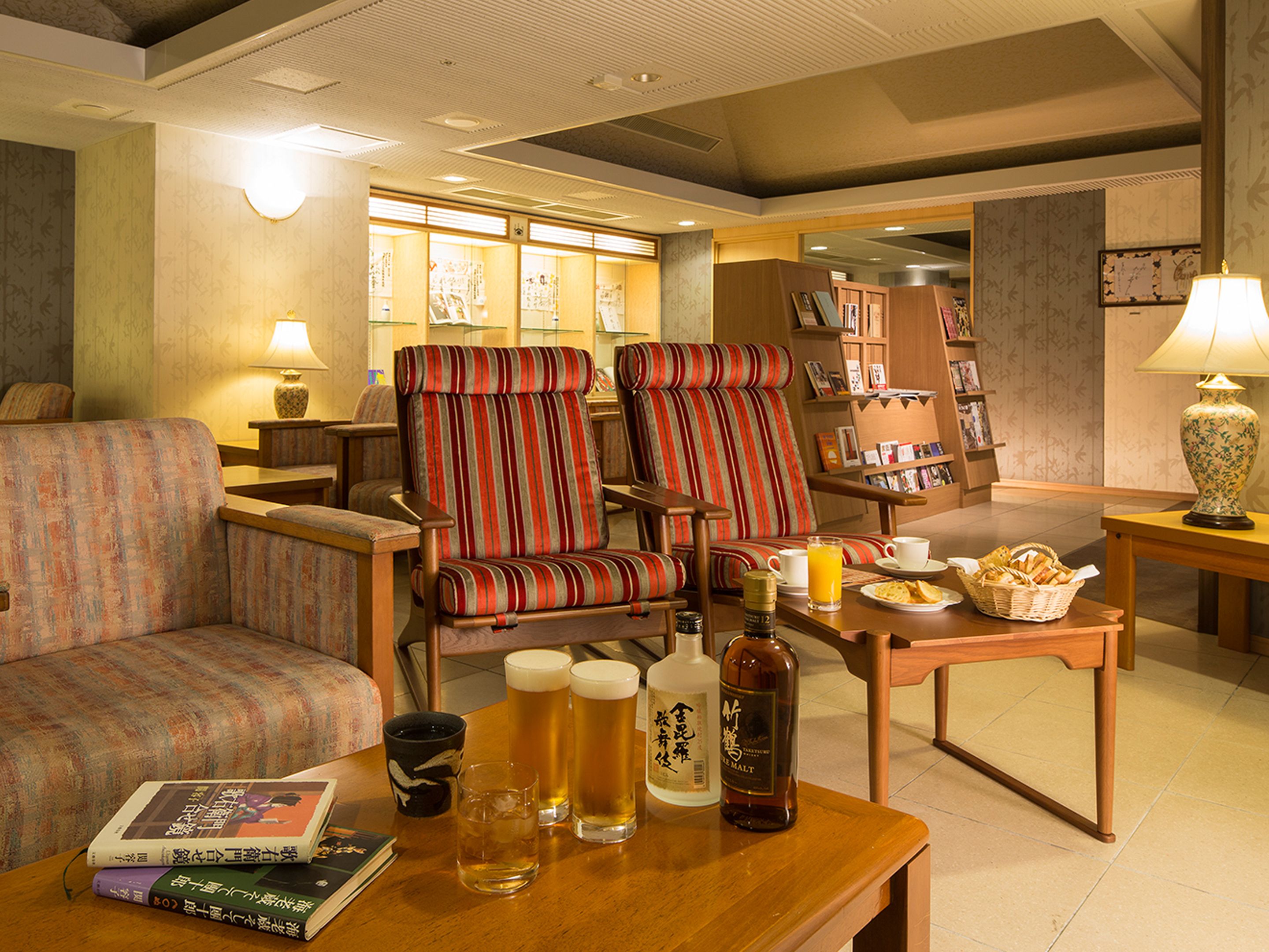 Ein gemütliches Hotelzimmer mit einer Couch, zwei gestreifte Sessel, einem Holztisch mit Getränken und Snacks sowie einem Bücherregal im Hintergrund.