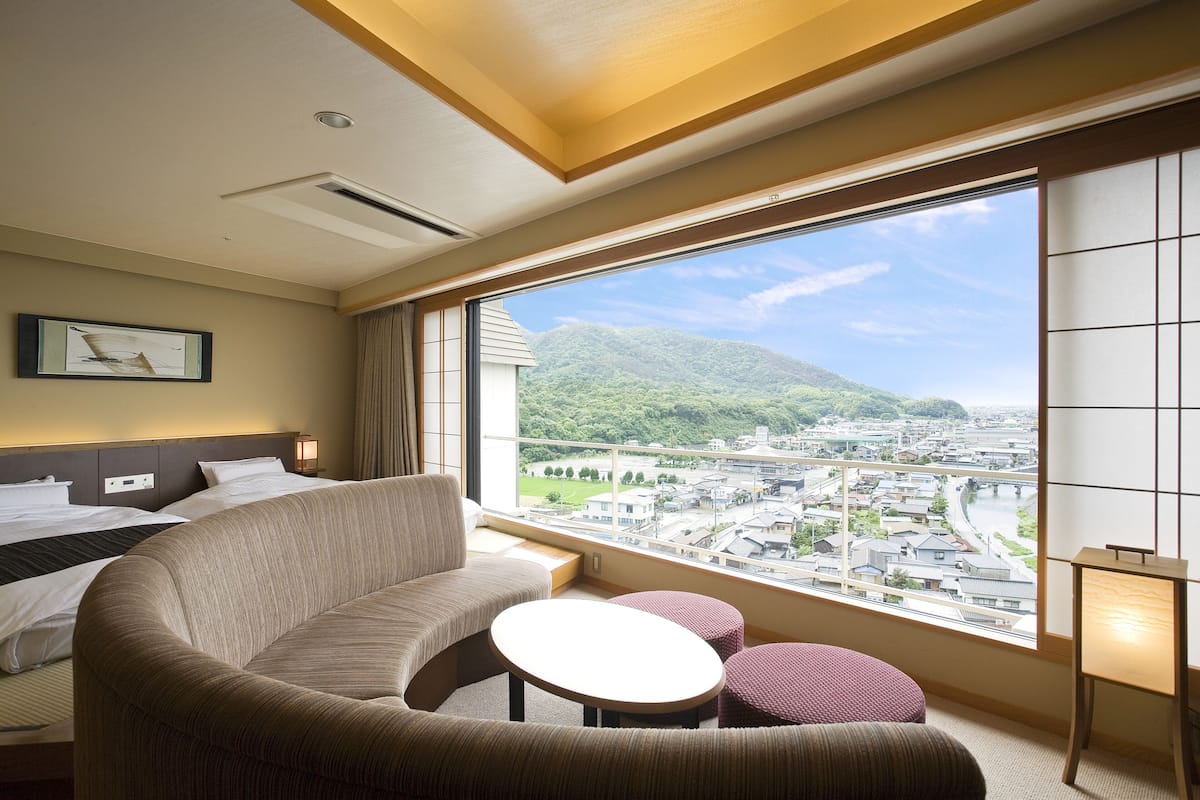 Une chambre d’hôtel dotée d’une grande fenêtre donnant sur le paysage urbain, d’un lit, d’un canapé et d’une petite table.