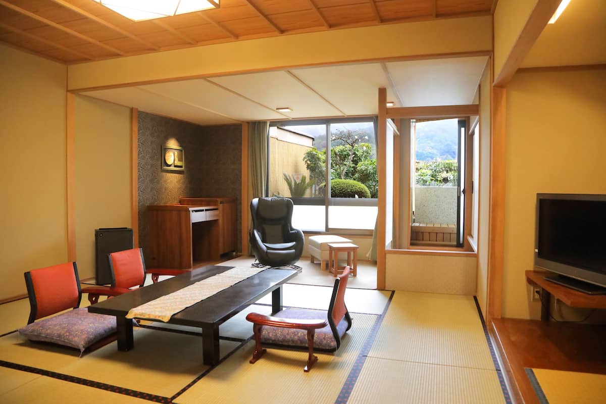 Une pièce de style japonais traditionnel, avec un sol recouvert de tatamis, une table basse, des chaises, un téléviseur et une vue sur un jardin, accessible par une porte coulissante.