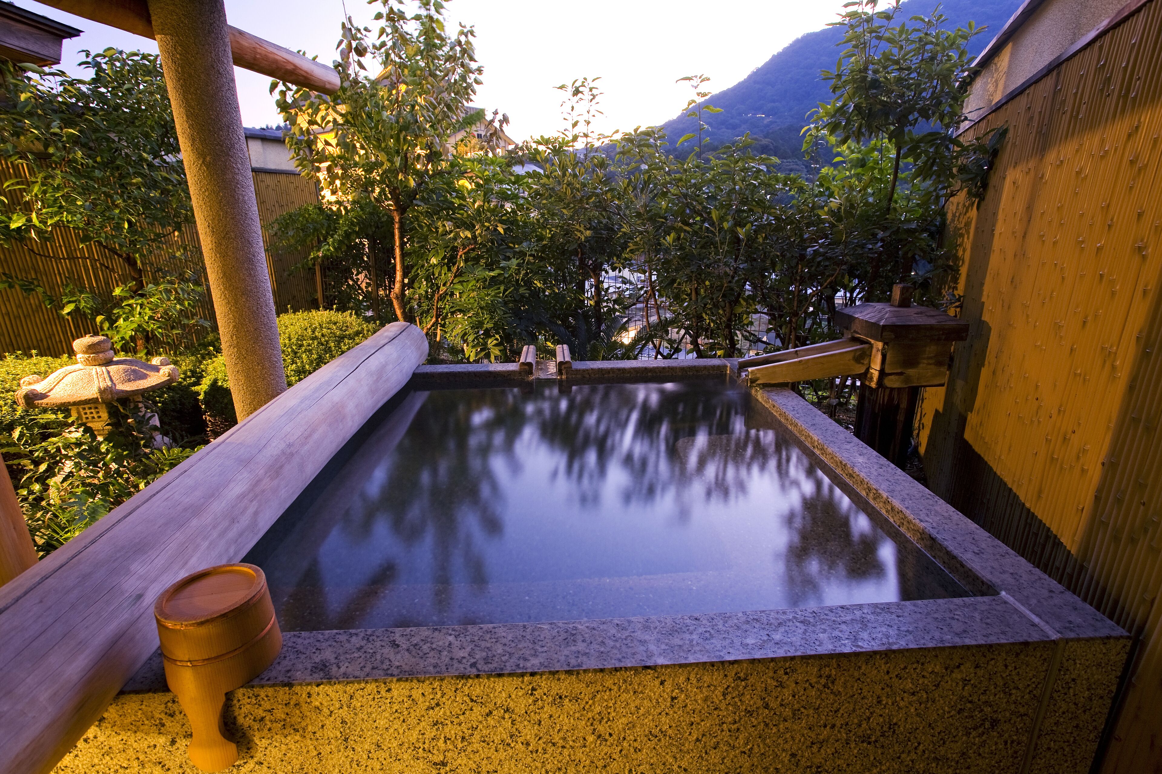 A traditional Japanese onsen with a wooden walkway leading to a rectangular hot spring area, surrounded by bamboo and trees.