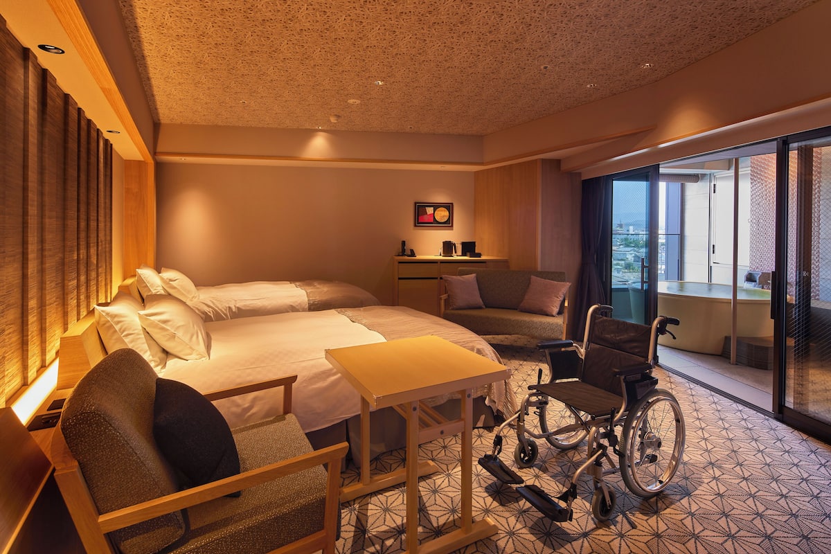Une chambre d’hôtel équipée d’un lit, d’un fauteuil roulant, d’un bureau et offrant une vue sur la ville grâce à de grandes fenêtres.