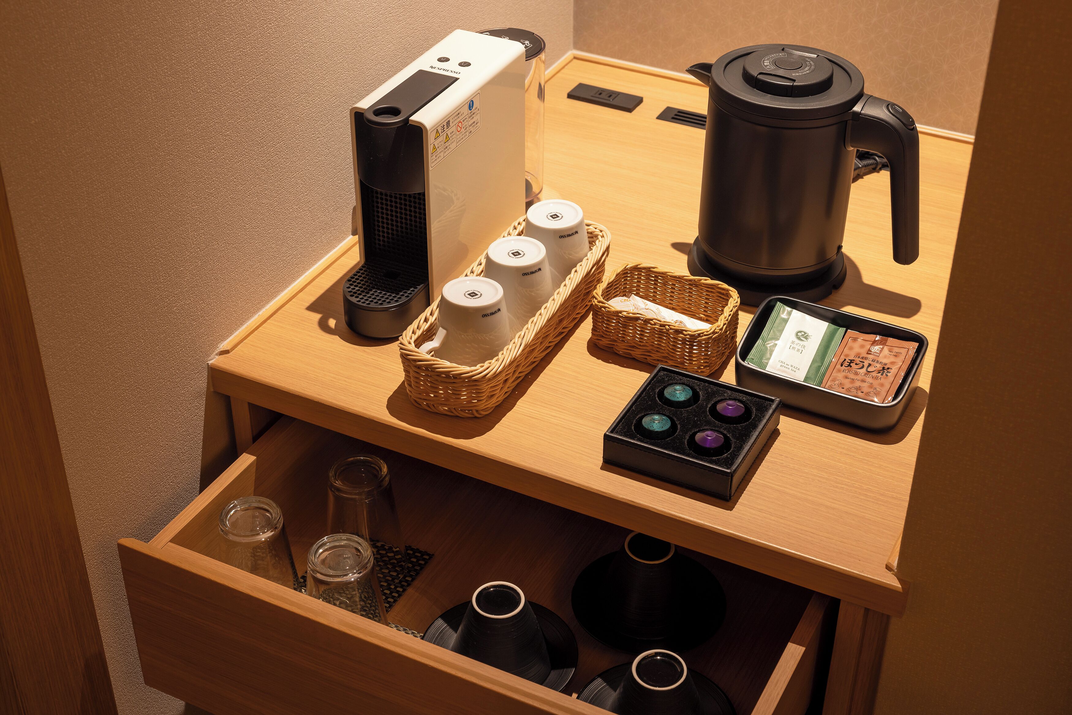 A wooden table with a coffee maker, a kettle, a basket with cups, a small device, and a drawer with tea pots.