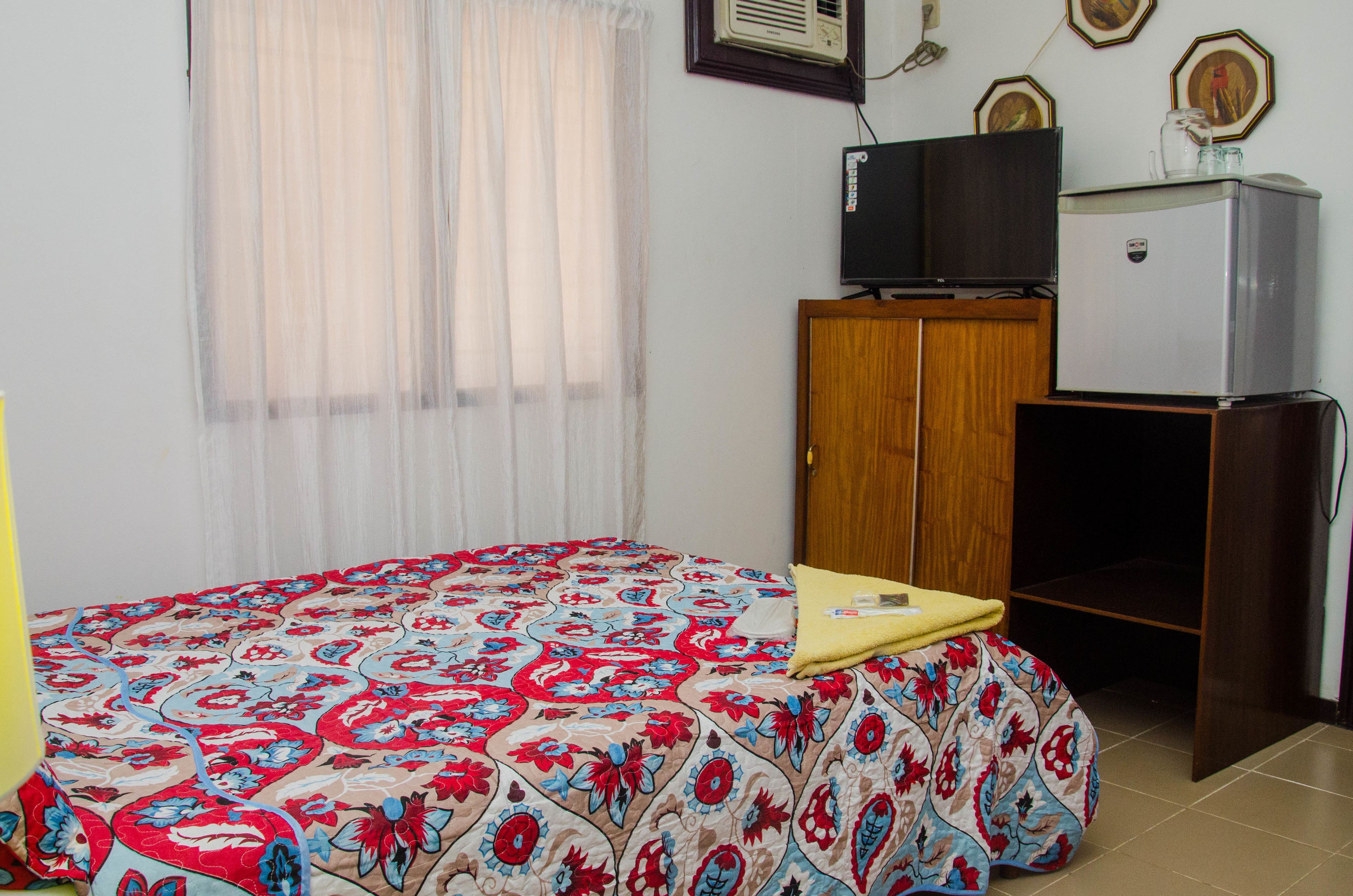 Un dormitorio con cama, un armario de madera, un refrigerador pequeño y un televisor sobre un mueble de madera.