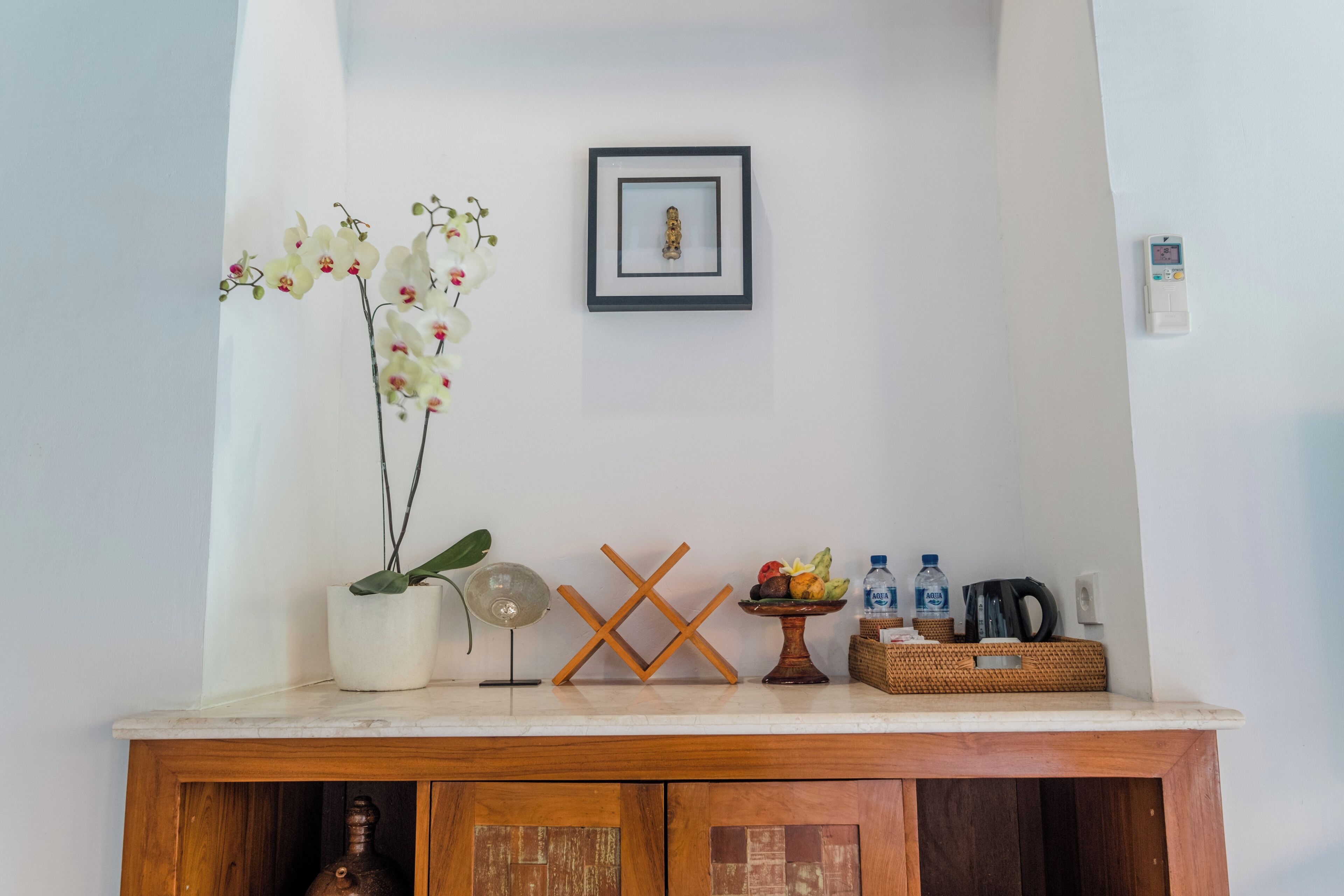 Un mueble de madera con una orquídea blanca, un cuadro enmarcado, un pequeño ventilador, un frutero y una botella de agua.