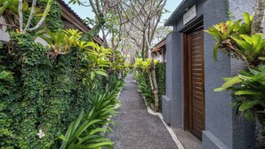 A narrow alleyway with a pathway flanked by lush greenery and a building with a wooden door. - Alam Bidadari Seminyak Villas (Seminyak)