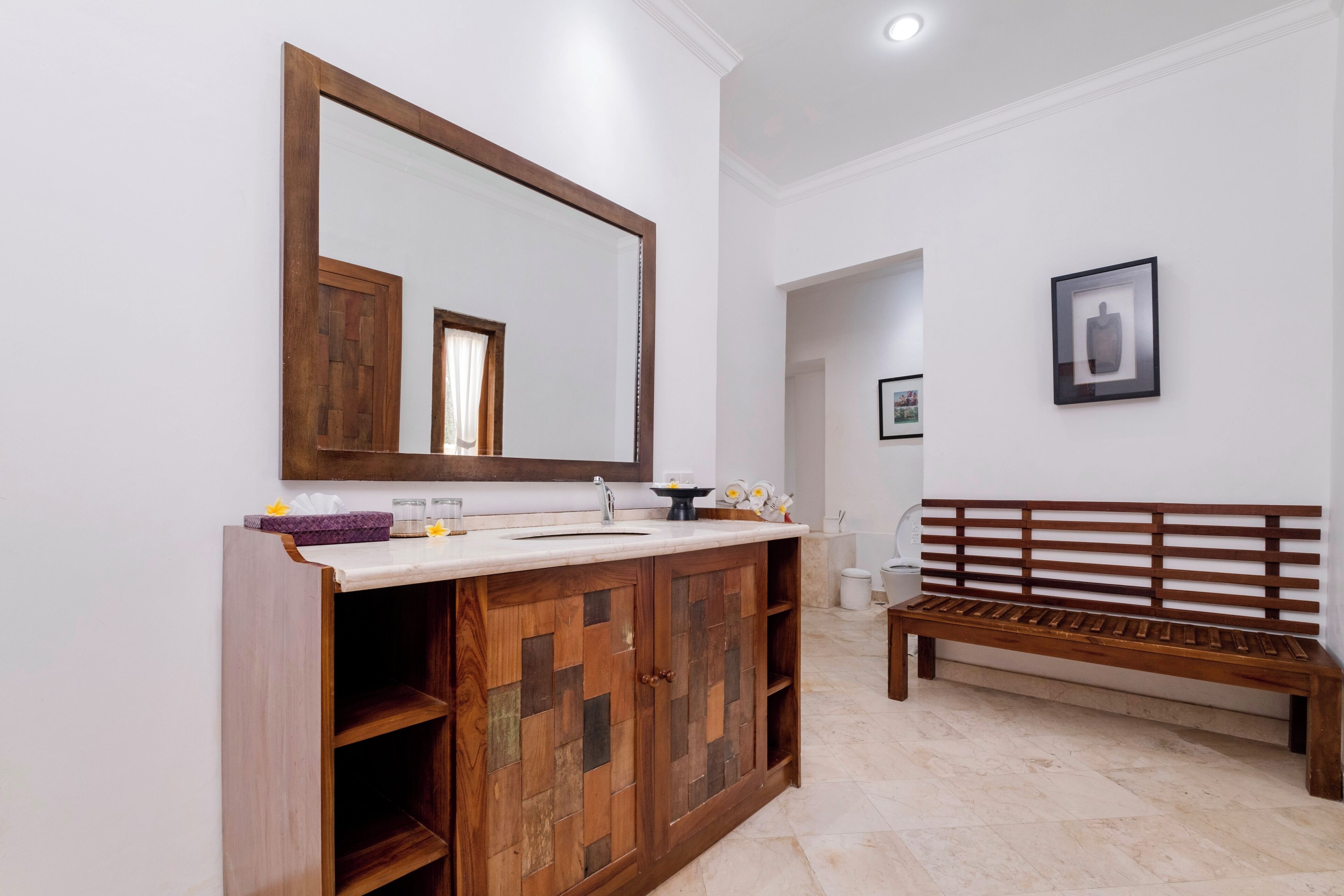 A bathroom with a wooden cabinet, a large mirror, a bench, and a framed picture on the wall.