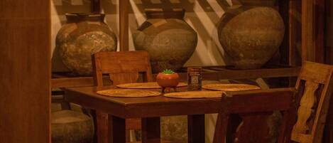A wooden table with woven placemats, a small potted plant, and a QR code. Behind the table, there are shelves with various pottery items.