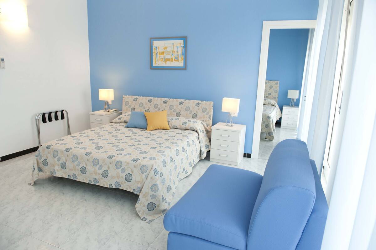 Un dormitorio con cama, mesitas de noche, lámpara, una silla azul y un espejo.