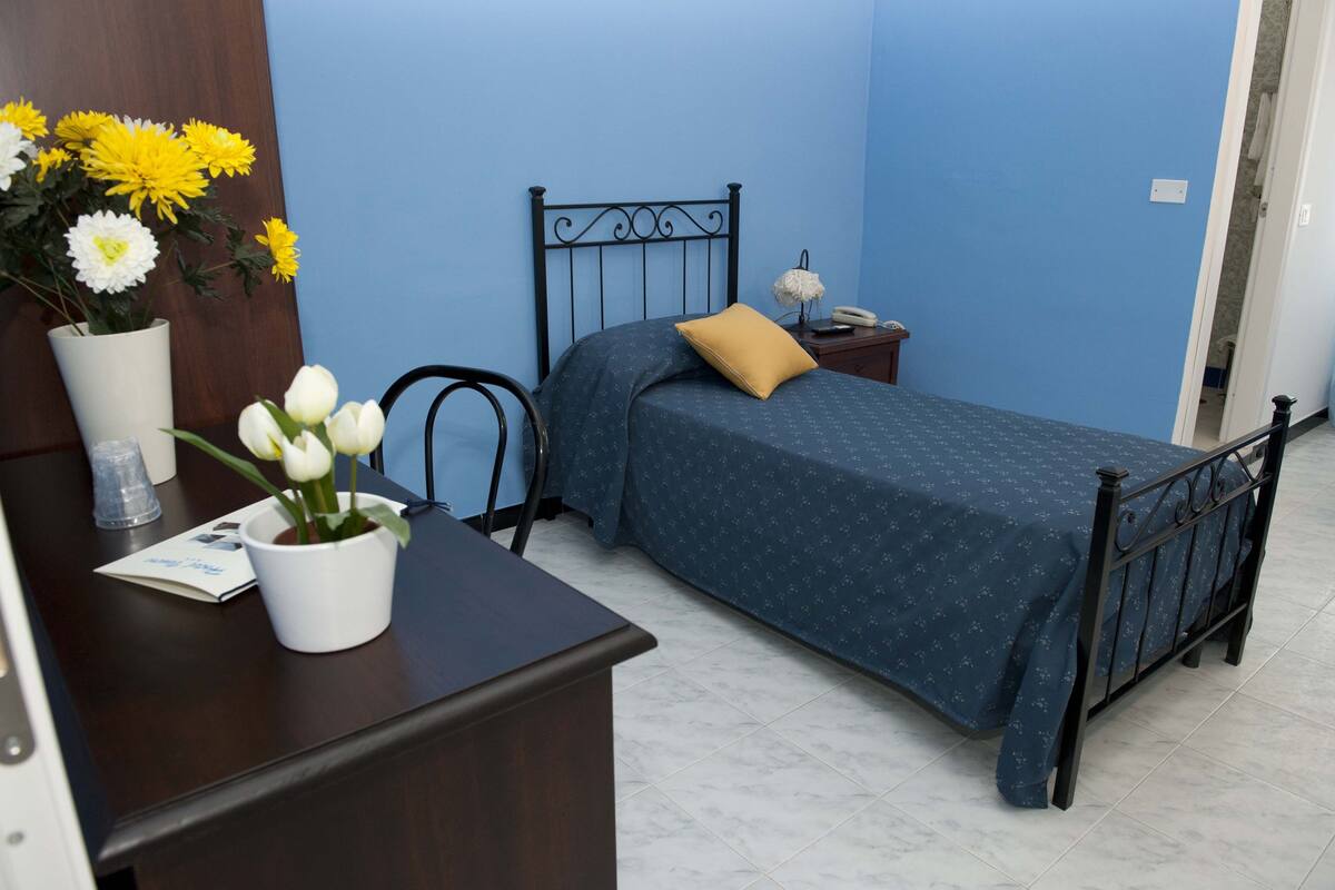 Un dormitorio con cama, mesita de noche, escritorio y silla. Hay flores sobre el escritorio y un jarrón con tulipanes.
