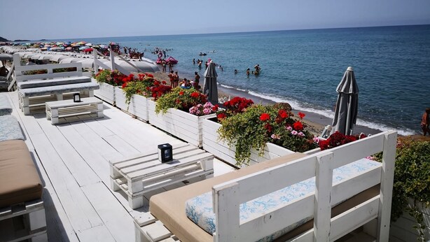 Une terrasse au bord de la plage, avec des bancs et des tables en bois peints en blanc, d’où l’on a une vue sur la mer, où l’on peut voir des gens nager et prendre des bains de soleil.
