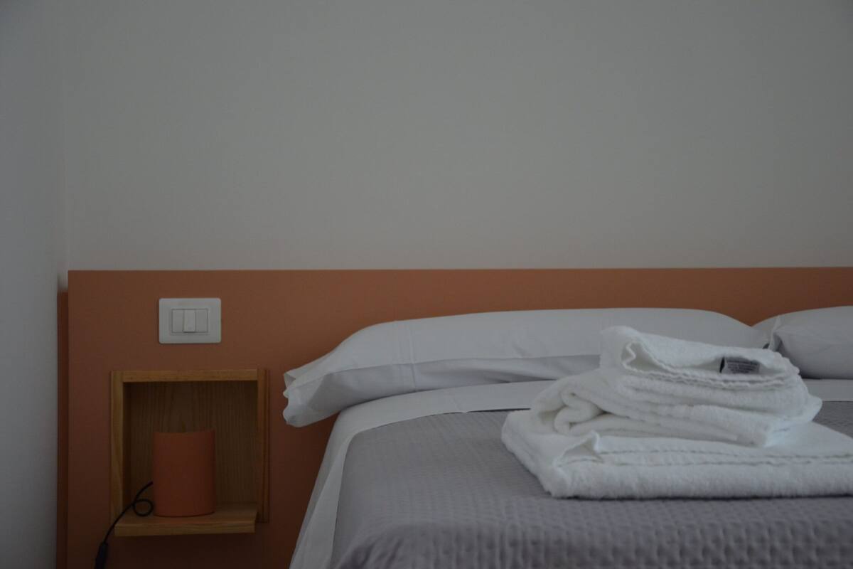 Una cama bien hecha con sábanas blancas, una mesita de noche de madera con una lámpara y un interruptor de luz en la pared.