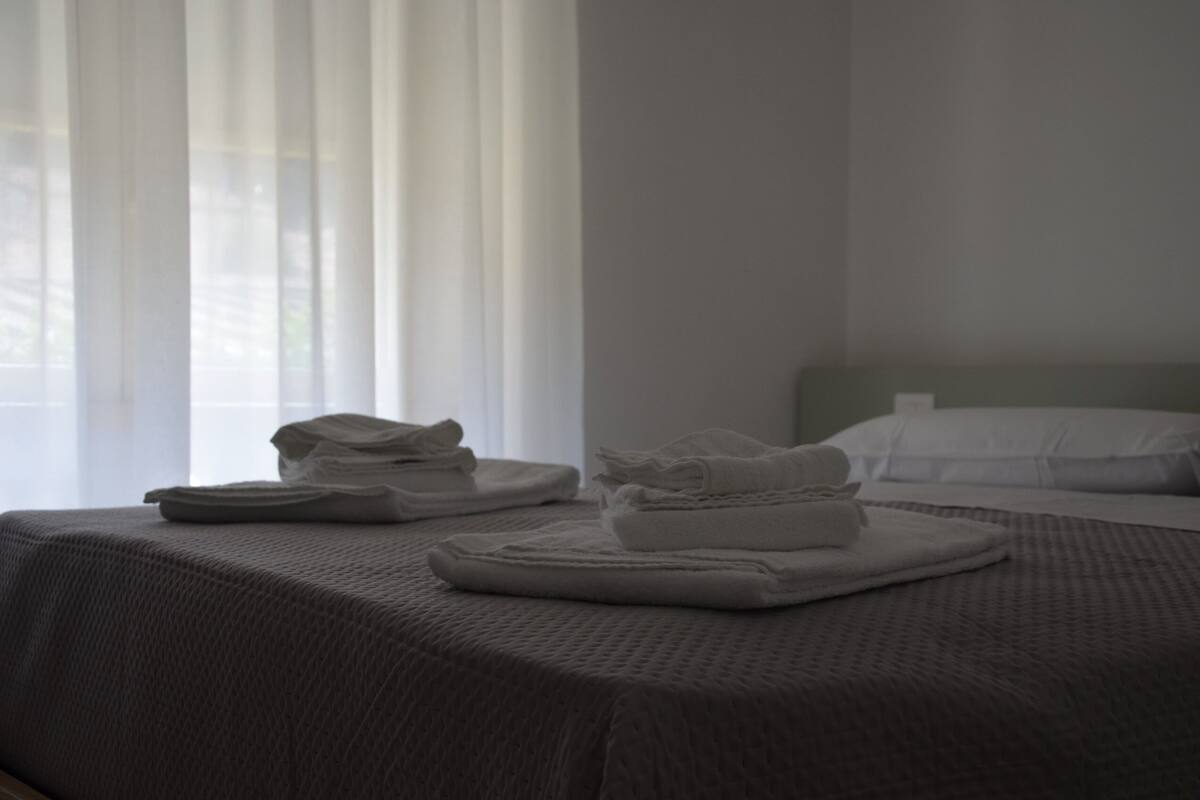 Habitación de hotel con dos camas, un ventanal con cortinas traslúcidas y toallas dobladas con cuidado sobre las camas.