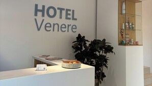 Un comptoir d’accueil avec une enseigne indiquant « HÔTEL Venere ». Il y a une plante sur la droite et une étagère avec des articles de toilette.
