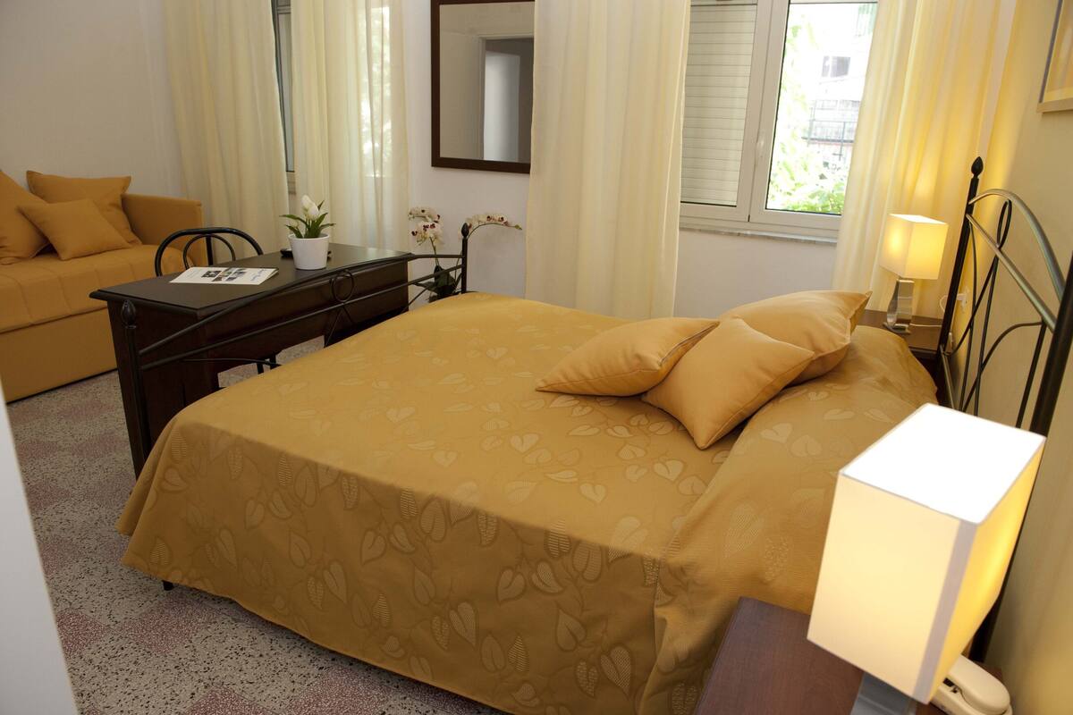 Una habitación de hotel con cama, escritorio, silla, lámpara y ventana con cortinas.