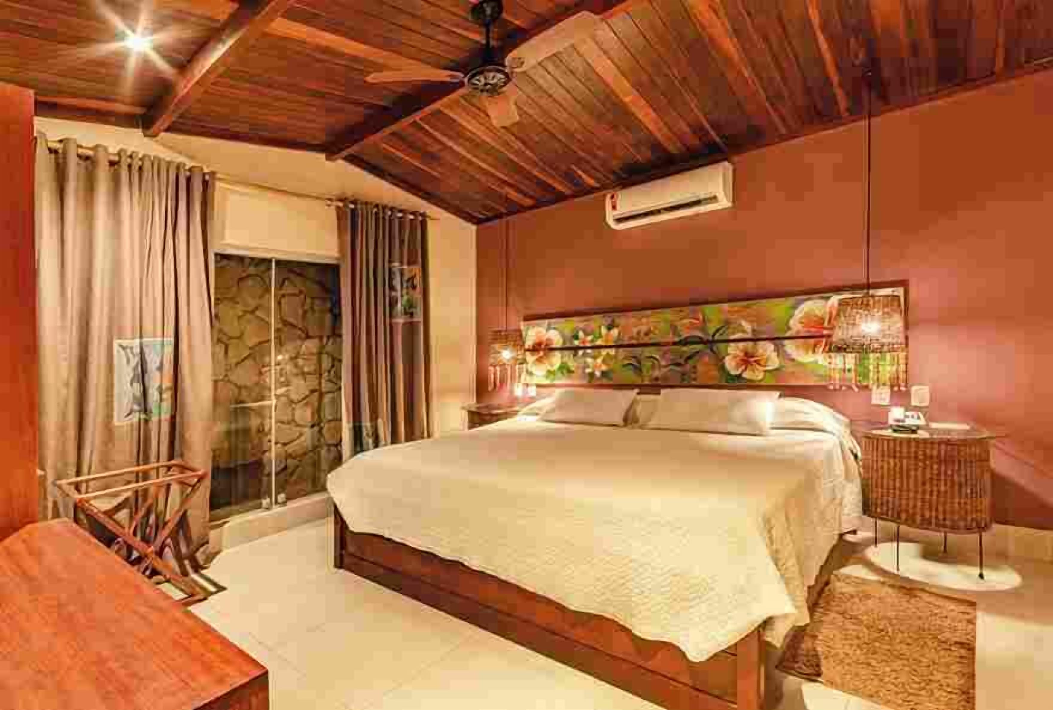 A bedroom with a wooden ceiling, a bed with a floral headboard, a nightstand, and a window with curtains.