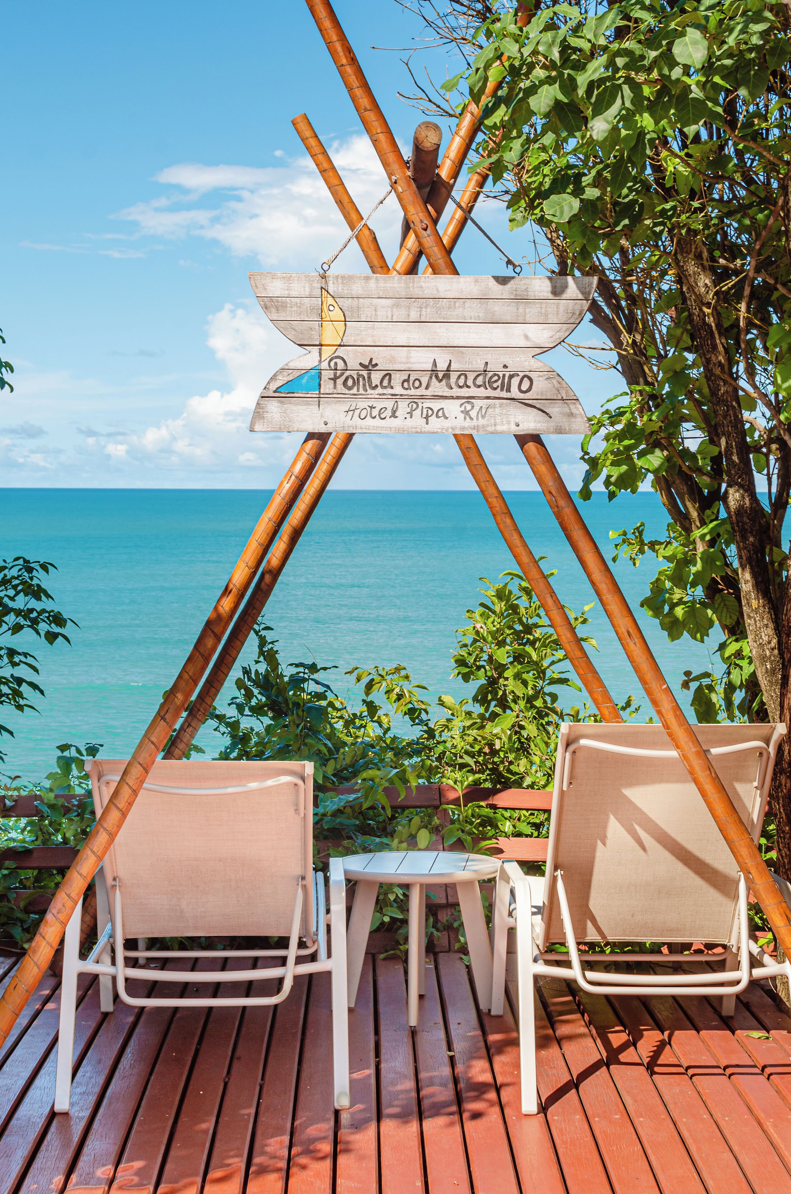 Une enseigne en bois portant les inscriptions « Ponta do Madeira Hotel » et « Pipa RN », placée devant une terrasse en bois, deux chaises longues blanches, et offrant une vue sur l’océan.