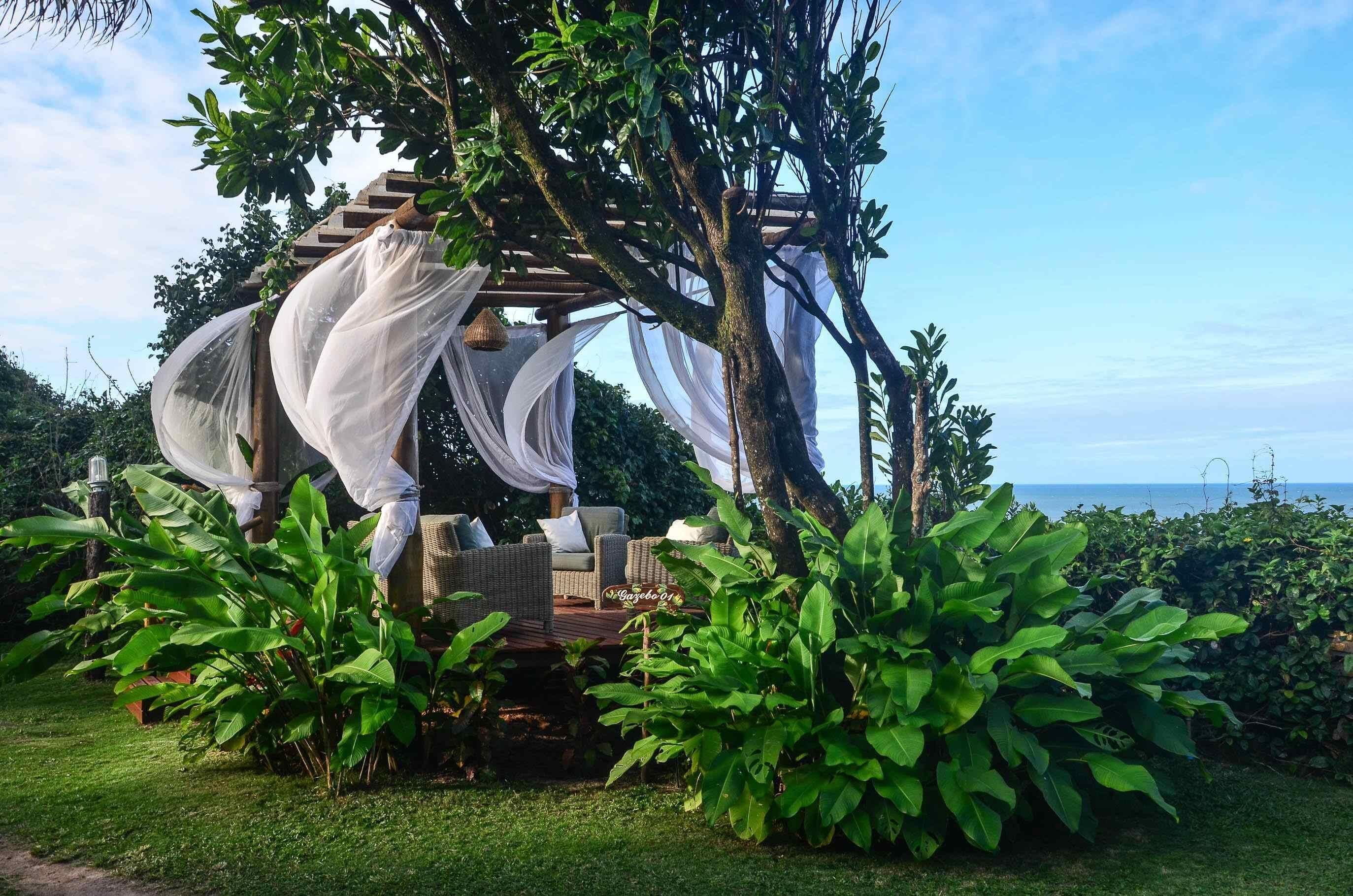 A gazebo with a wooden structure and white drapes, surrounded by lush greenery and a view of the sea.