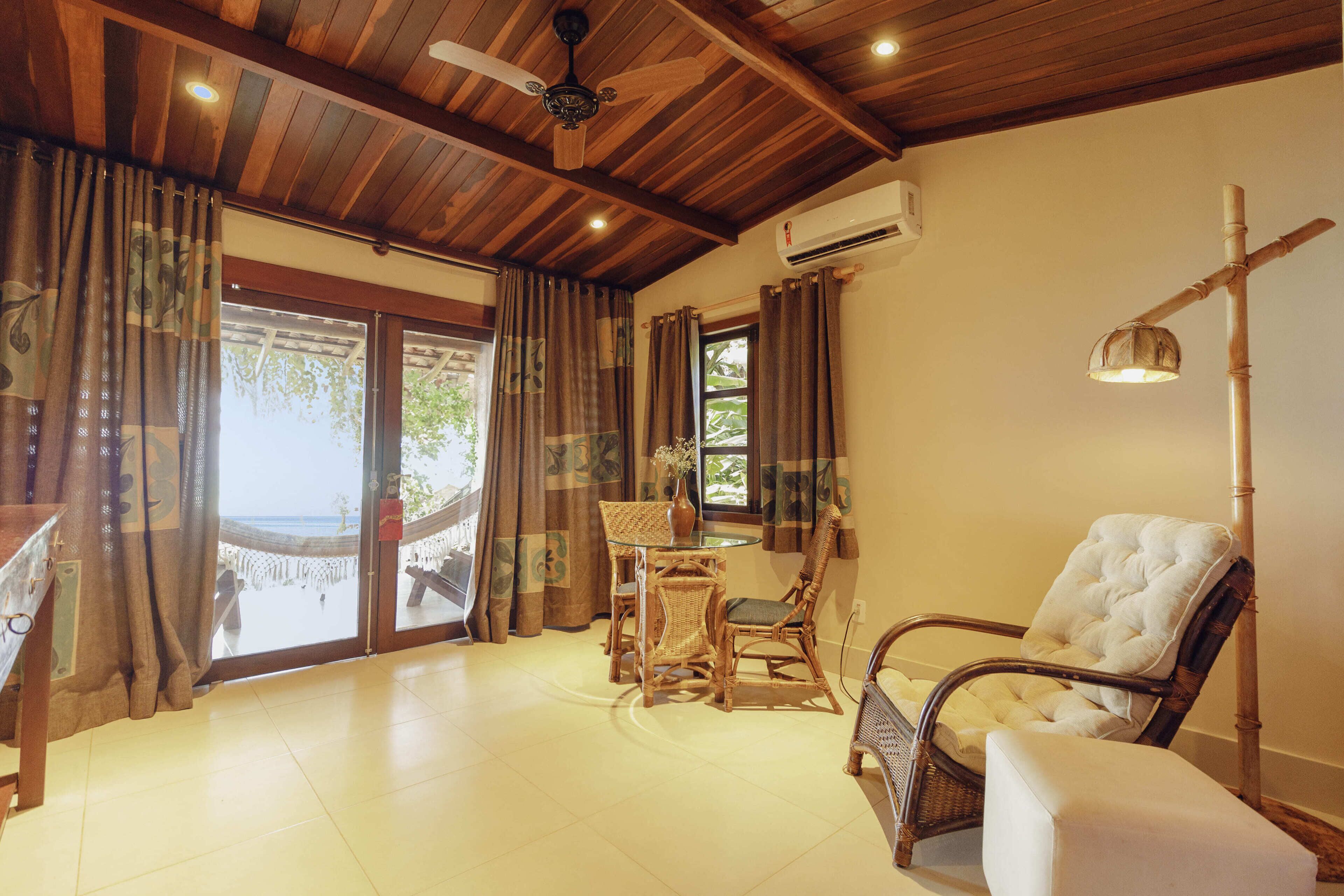 A room with a wooden ceiling, a glass table with chairs, a white cushioned chair, and a view of the beach through large windows.