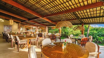 Un área de comedor al aire libre con mesas y sillas de madera, techo cubierto y exuberante follaje de fondo.