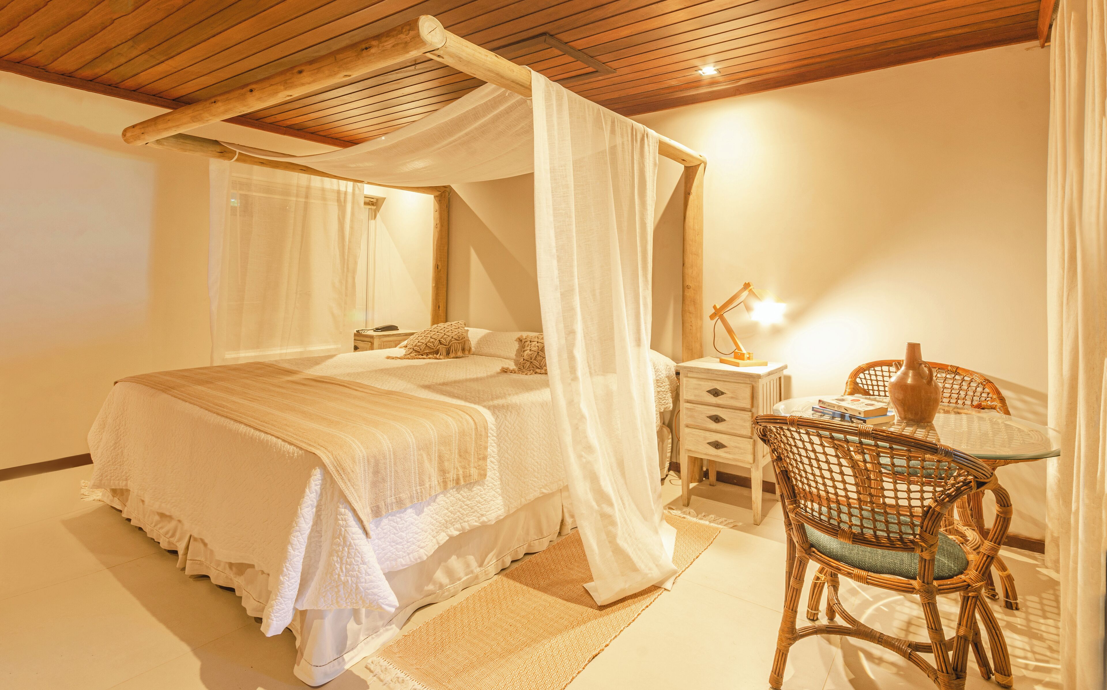 A bedroom with a four-poster bed, a bedside table with a lamp, and a wicker chair.
