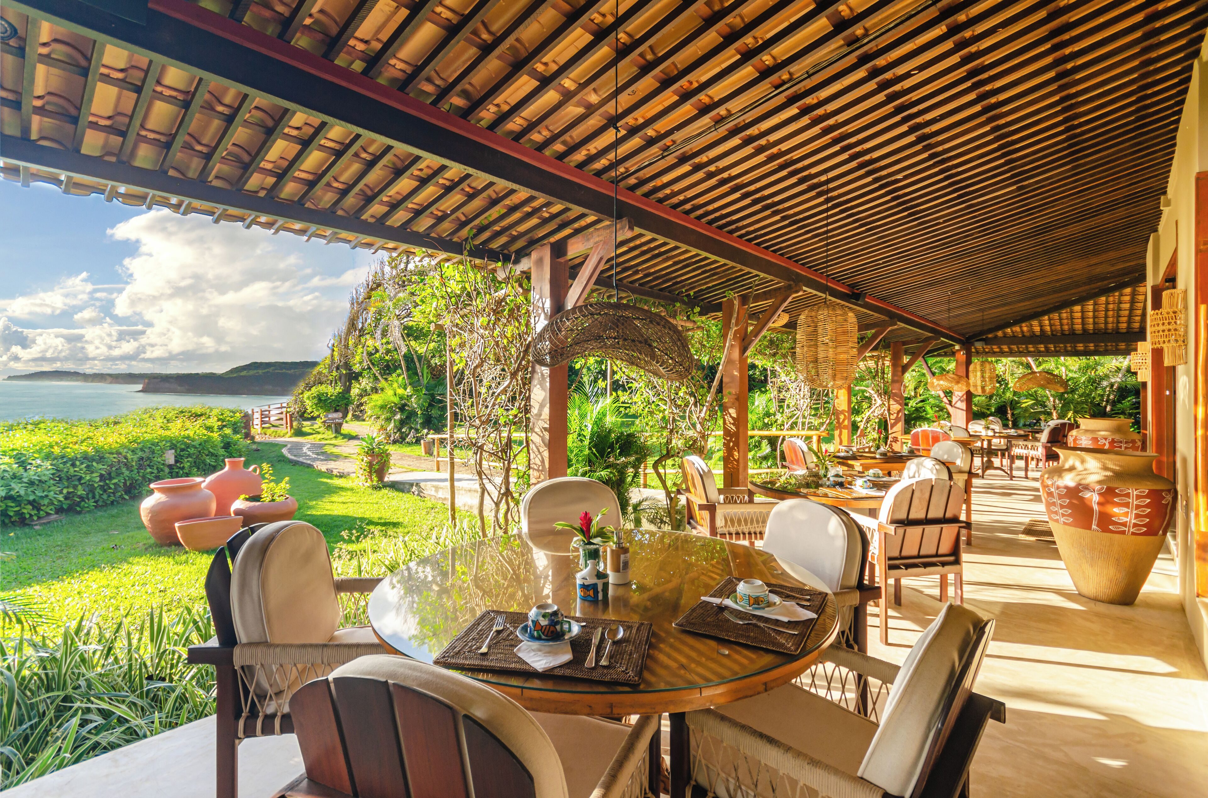 Un área de comedor al aire libre techada, con una mesa dispuesta para dos, rodeada de sillas, y vistas a un jardín exuberante y la costa.