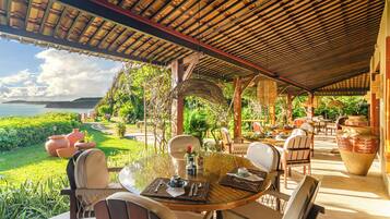 Un área de comedor al aire libre techada, con una mesa dispuesta para dos, rodeada de sillas, y vistas a un jardín exuberante y la costa.