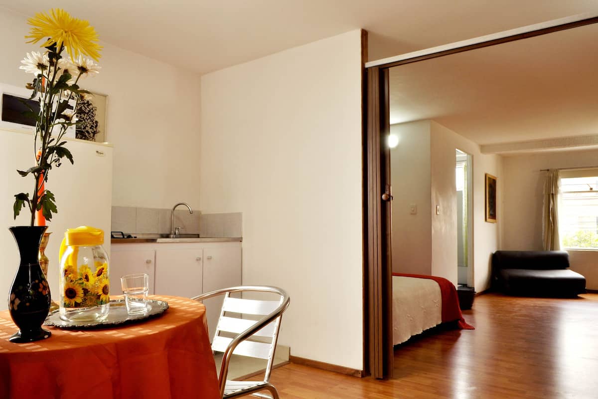 A room with a dining table, a chair, a vase with flowers, a glass of water, and a kitchen area with cabinets and a sink.